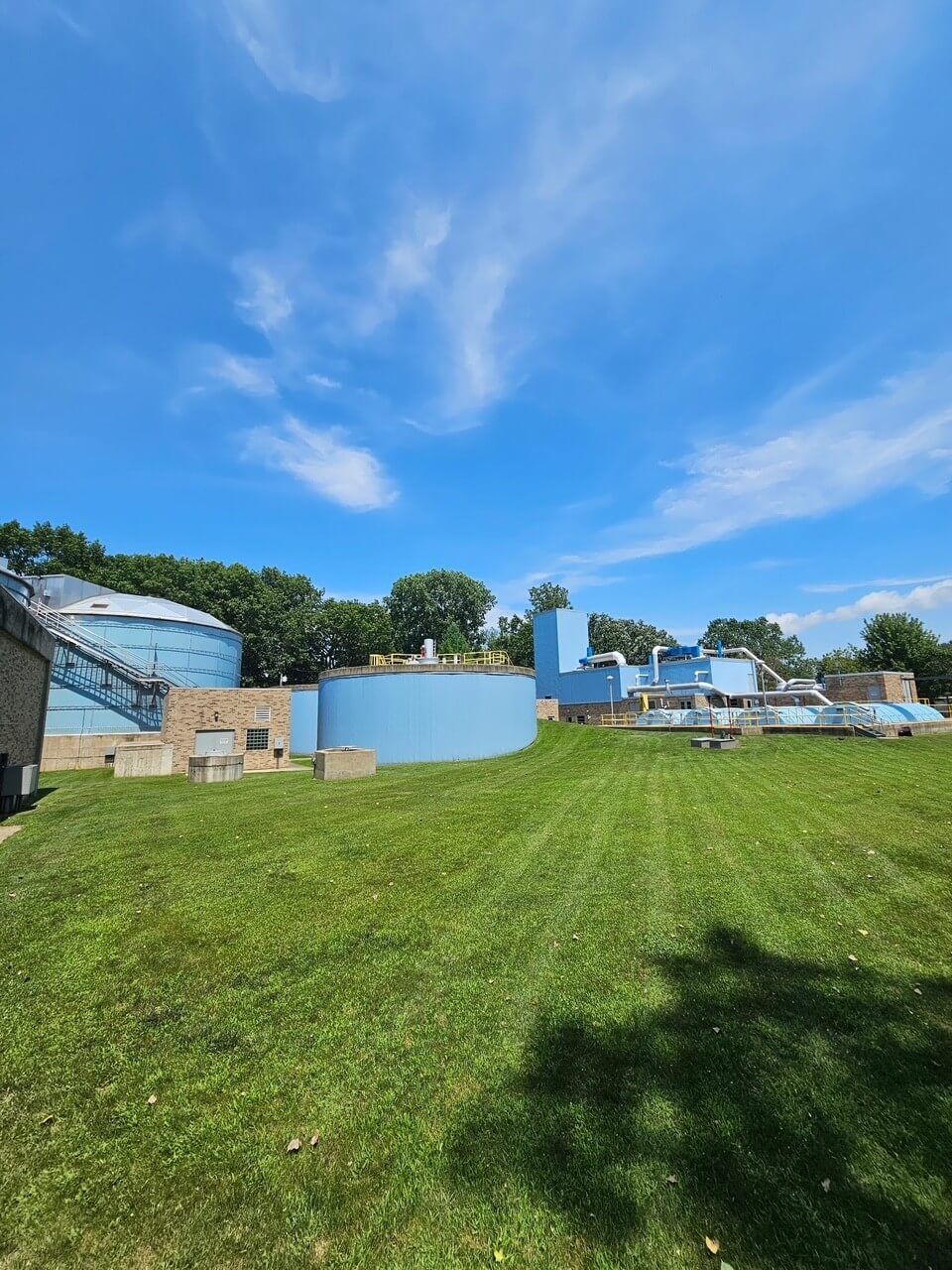 Grassy hill with a blue sky, trees, and a light-blue structure beside outdoor seating.
