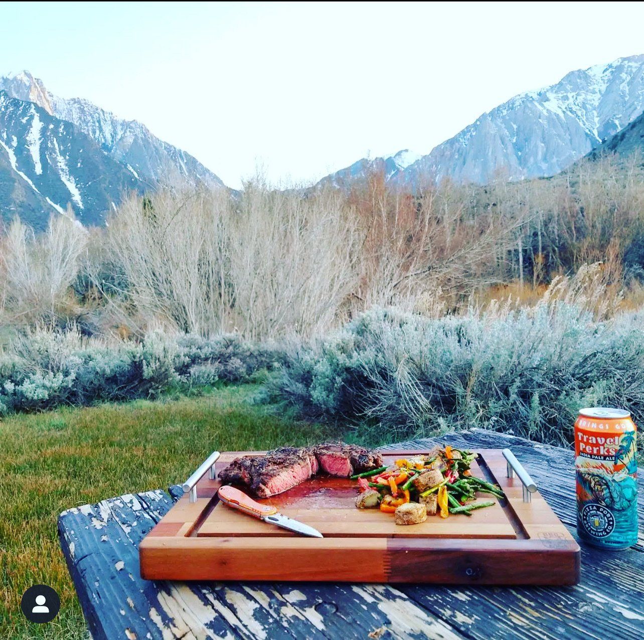 A steak and vegetables on a cutting board on a picnic table with mountains in the background.