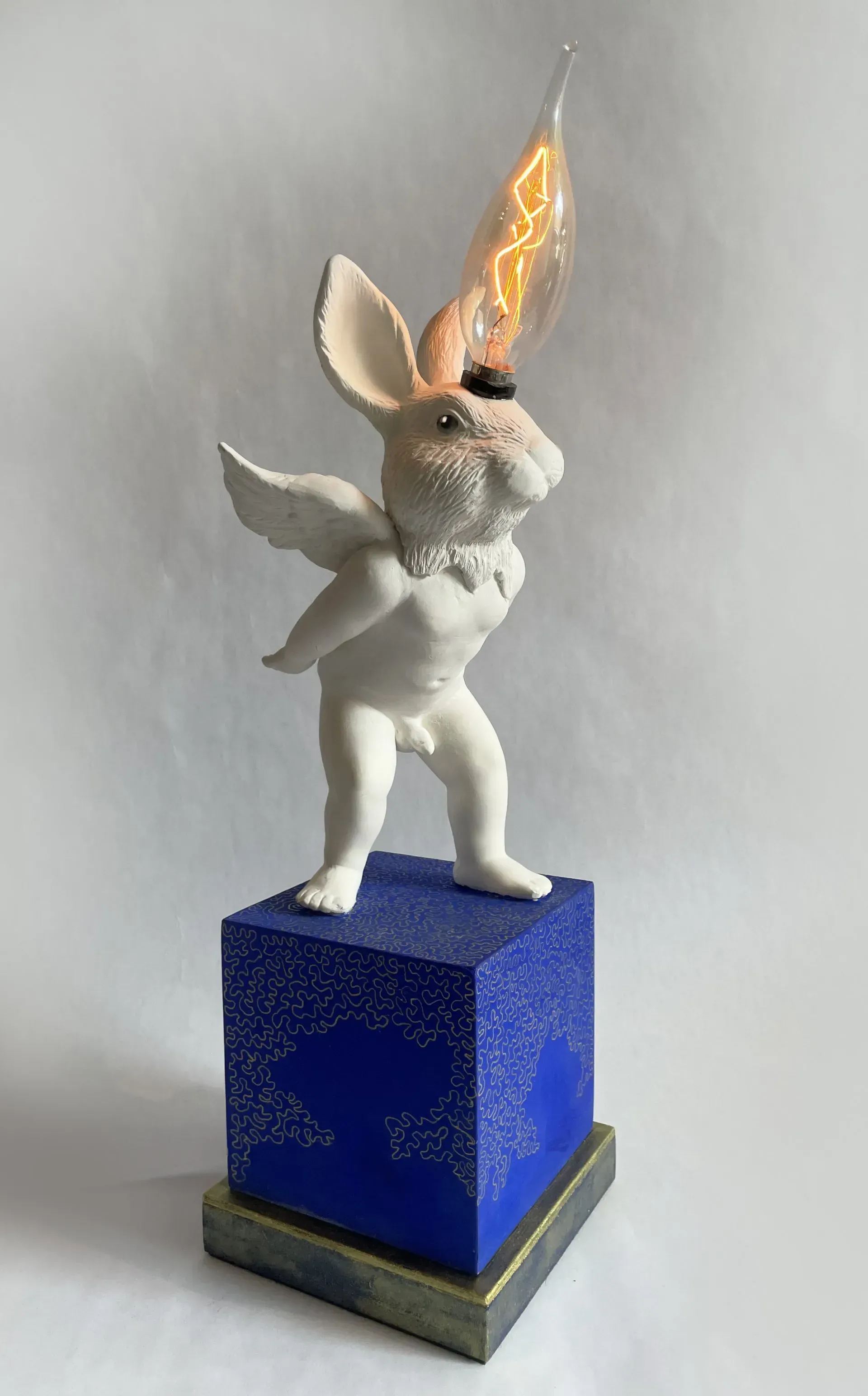 A whimsical rabbit sculpture with wings and a lightbulb head atop a blue pedestal.