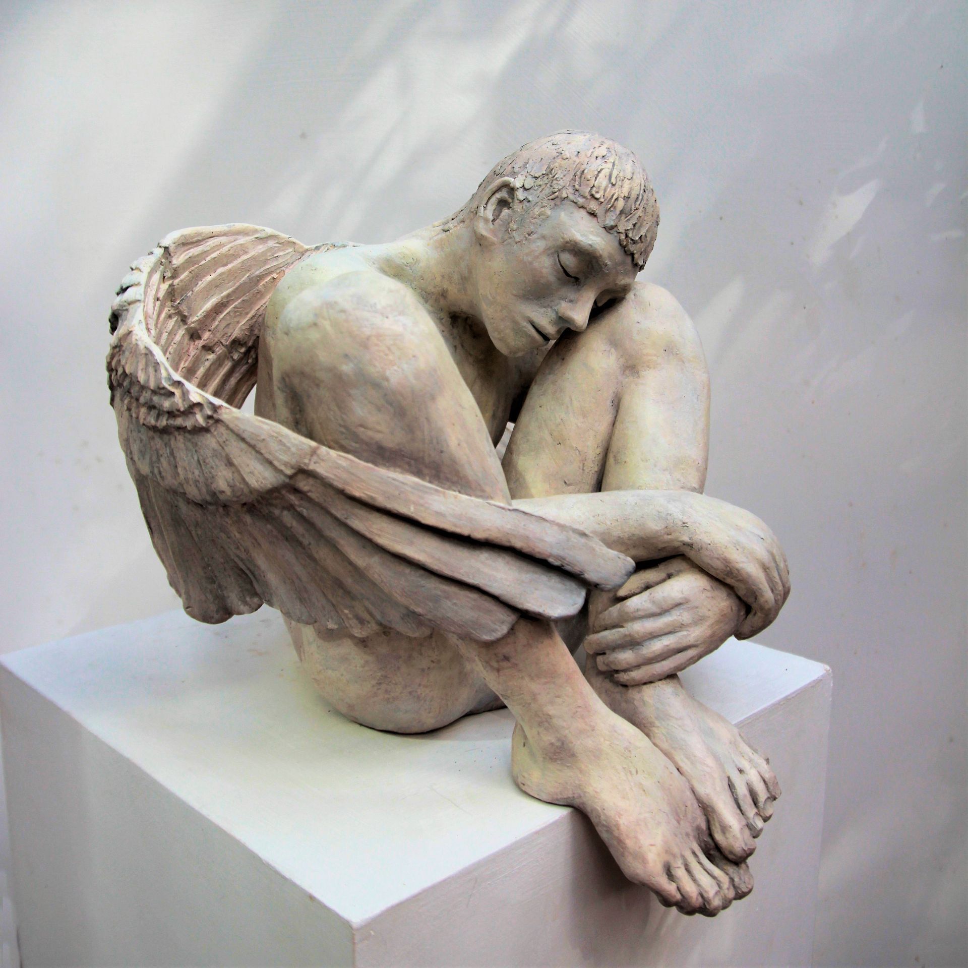 Sculpture of a winged figure, pale skin, seated, arms around knees, head down.by peter garrard