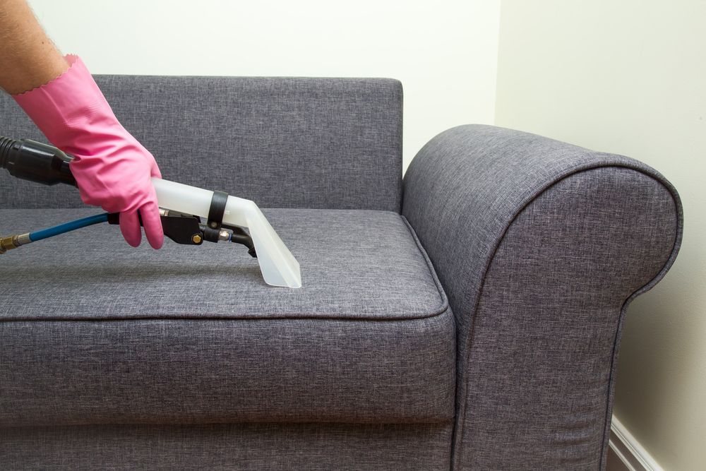 Person in Pink Gloves Cleaning a Gray Sofa With an Upholstery Cleaner — Kingaroy Carpet Cleaners in Nanango, QLD