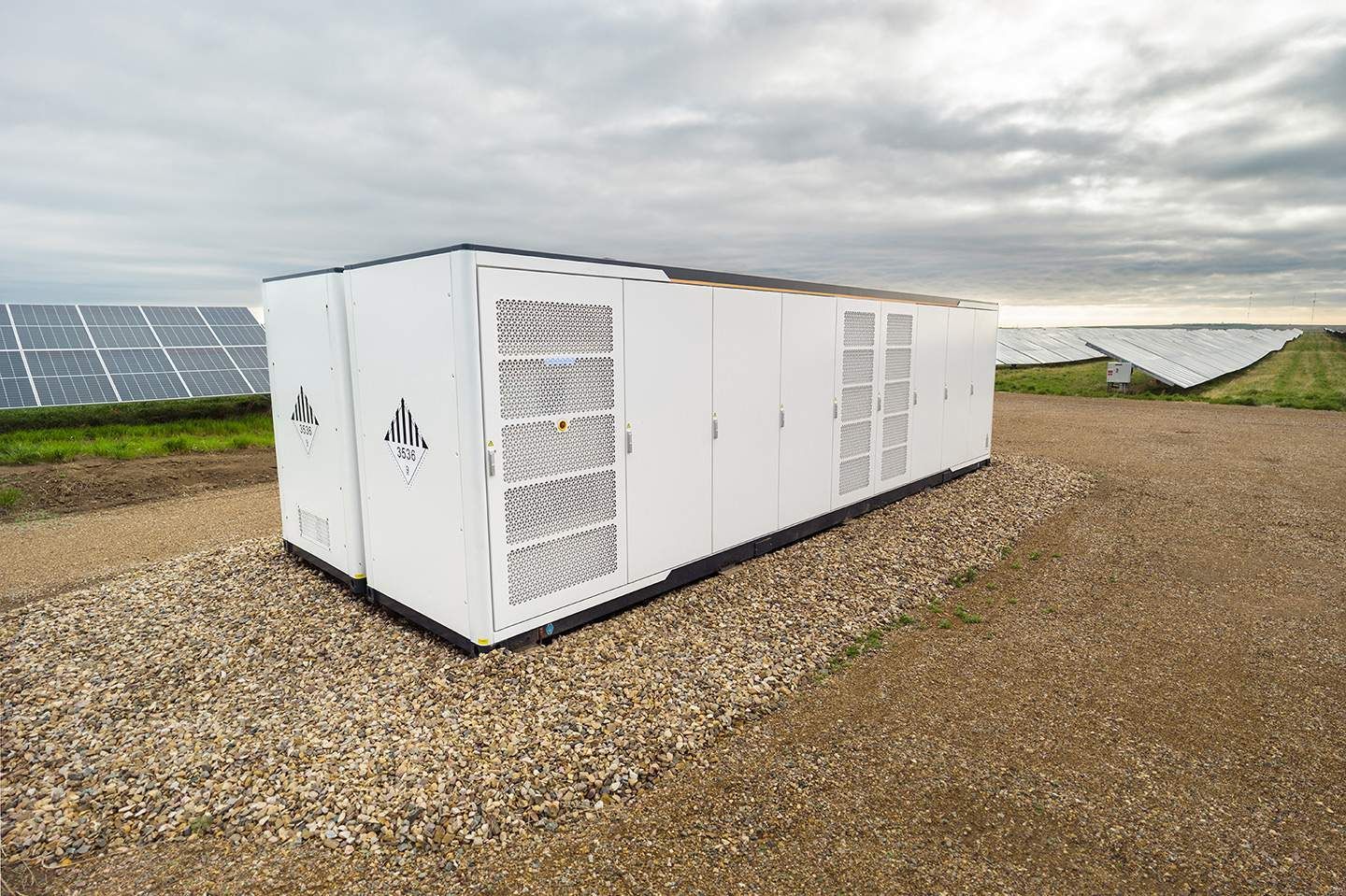 White energy storage unit with vent panels on gravel in front of solar panels under a cloudy sky.