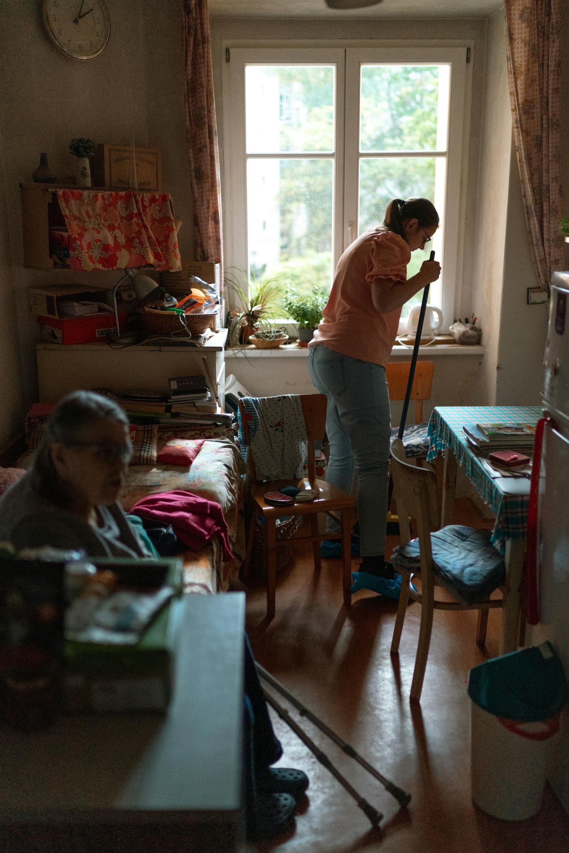 A person stands near a window holding a tool, while another person sits in the foreground of a cluttered home interior.