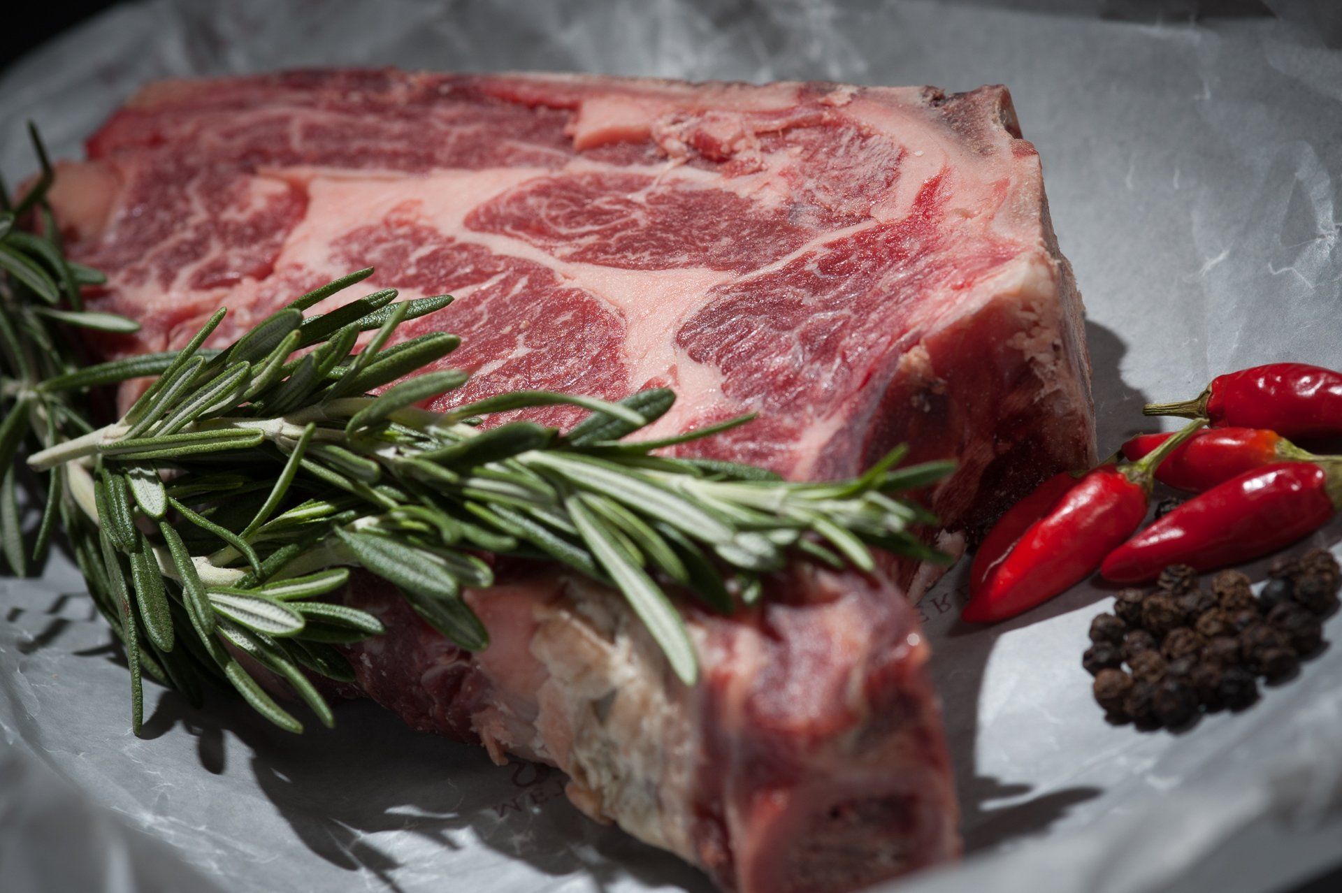 Bistecca di manzo cruda marmorizzata con rosmarino, peperoncino rosso e grani di pepe su carta da forno.