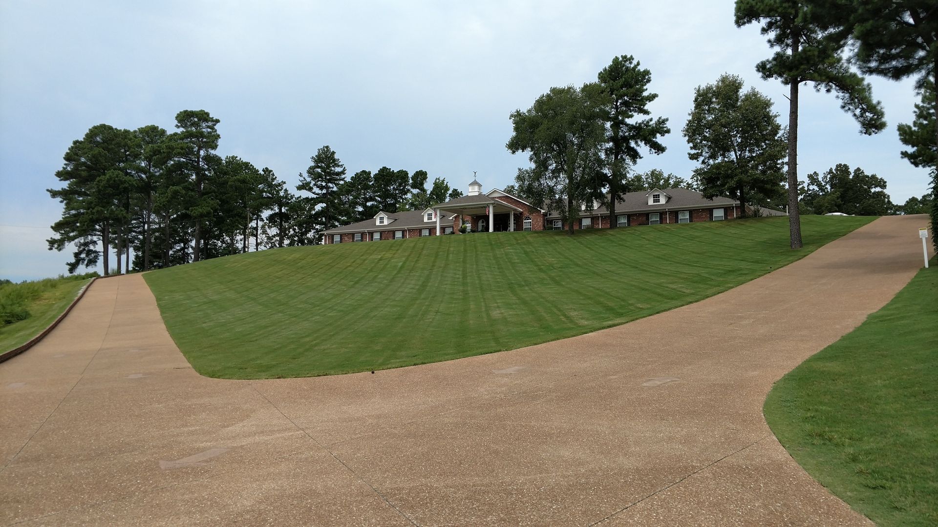 Gravel paths curve up a grassy hill toward a small building and trees under a cloudy sky.