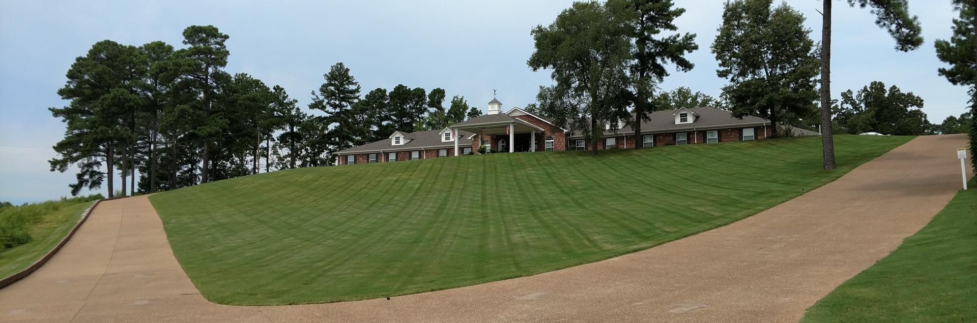 Curving gravel path through a green hill toward buildings and trees under a partly cloudy sky