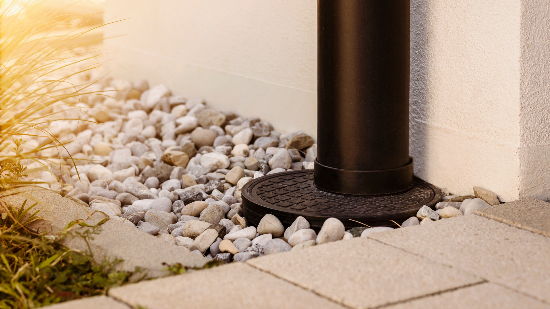 Black downspout drain on white gravel beside a beige wall and patio pavers