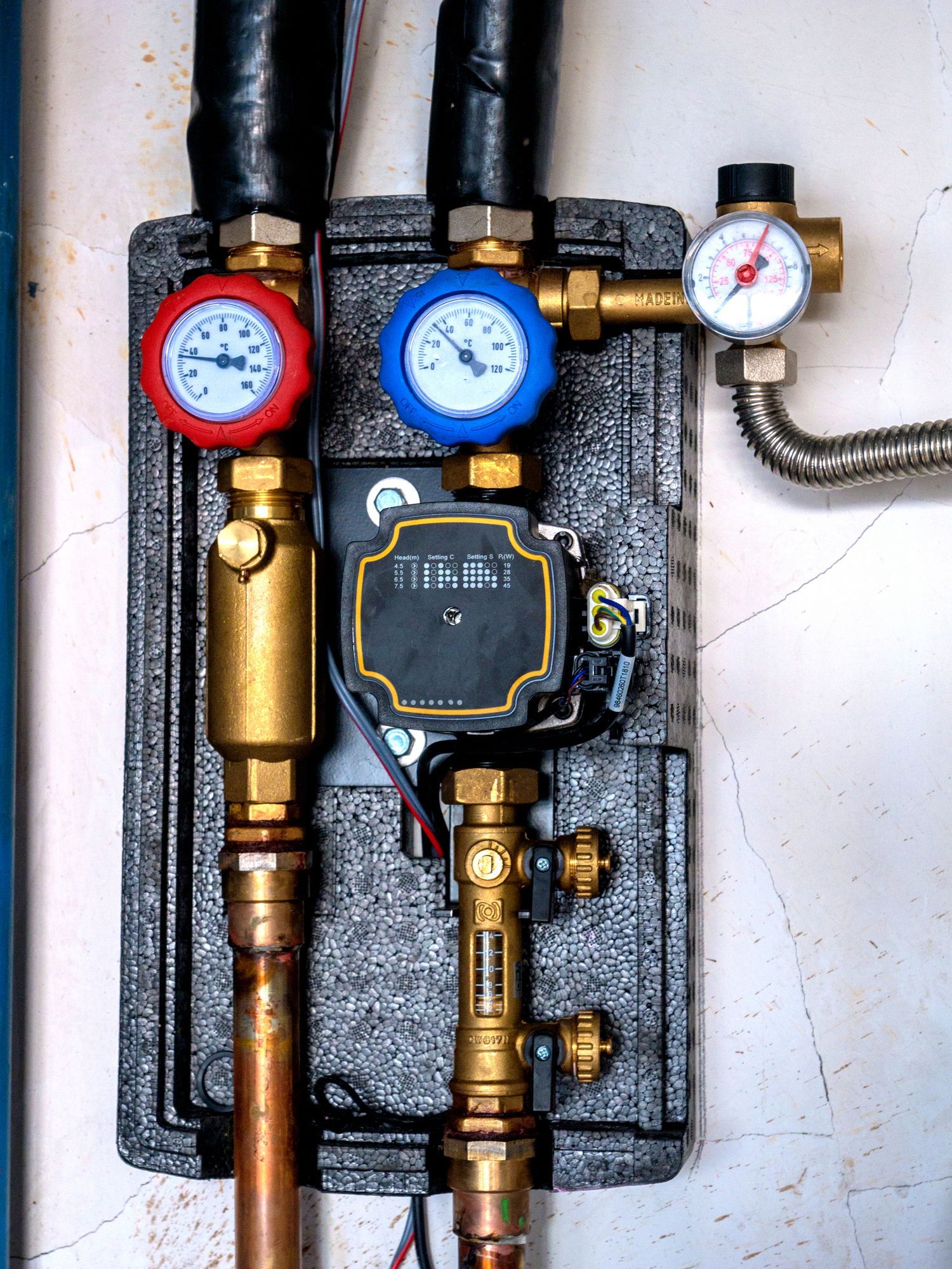 Close-up of a hydronic heating system with gauges, pump, and pipes against a wall.