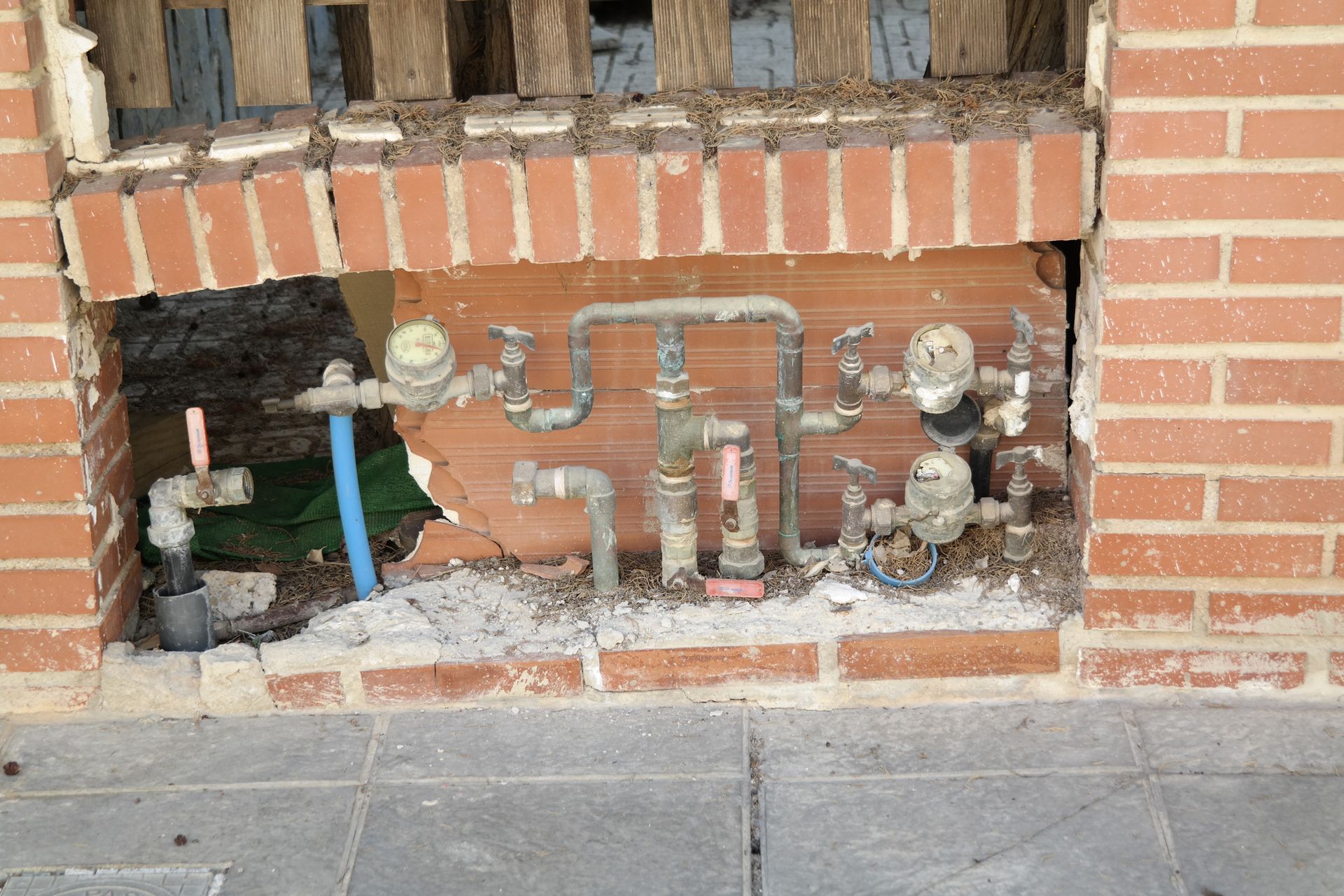Brick wall with exposed pipes and meters; damaged area reveals utility infrastructure.