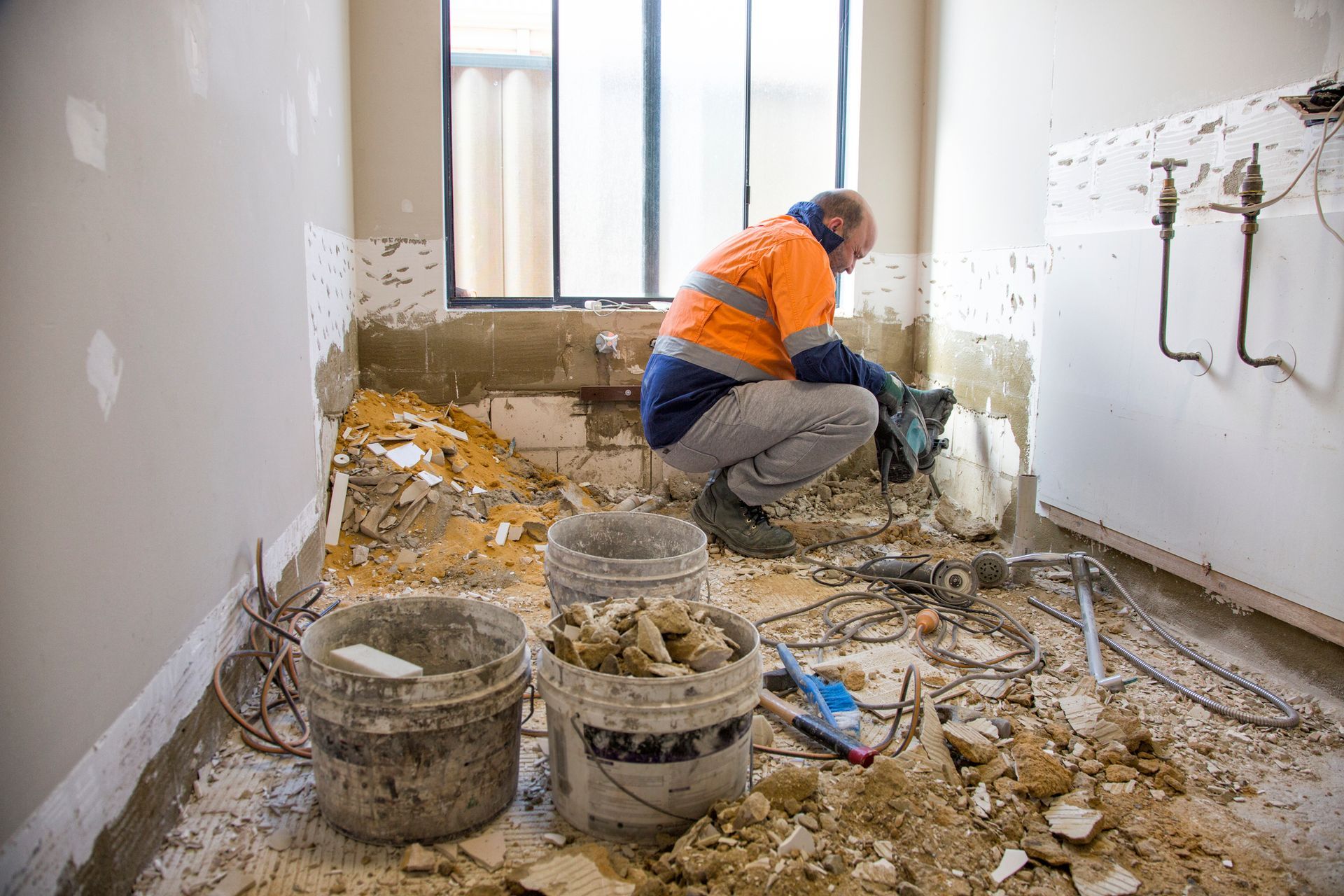 Construction worker demolishing a room with rubble and buckets. He wears an orange safety vest and kneels.