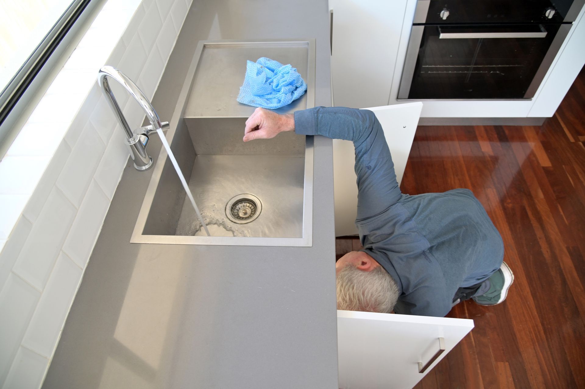 A person is under the kitchen sink, potentially repairing it. They are wearing a blue shirt.