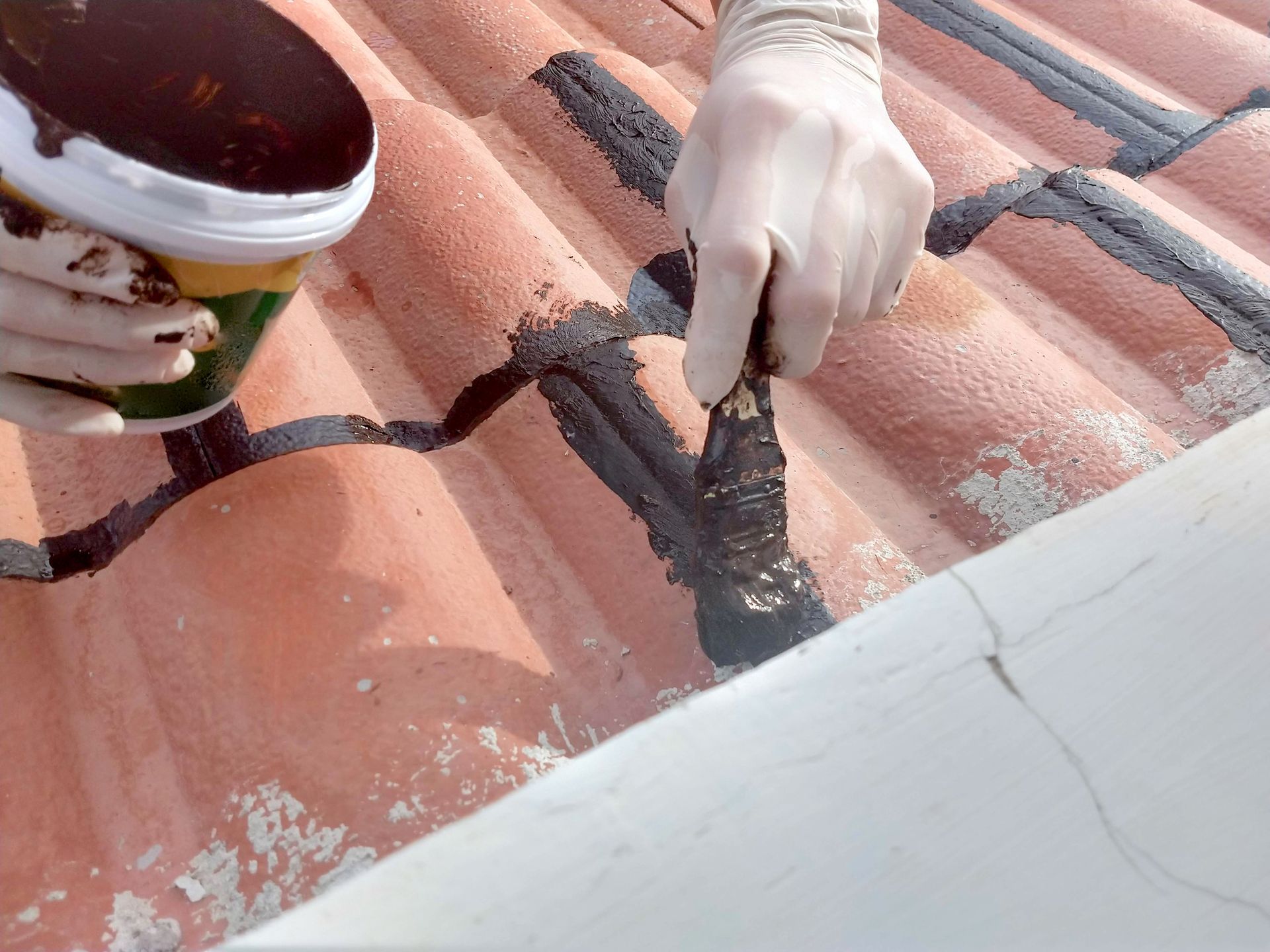 Person wearing gloves applying dark sealant to a terracotta tile roof.