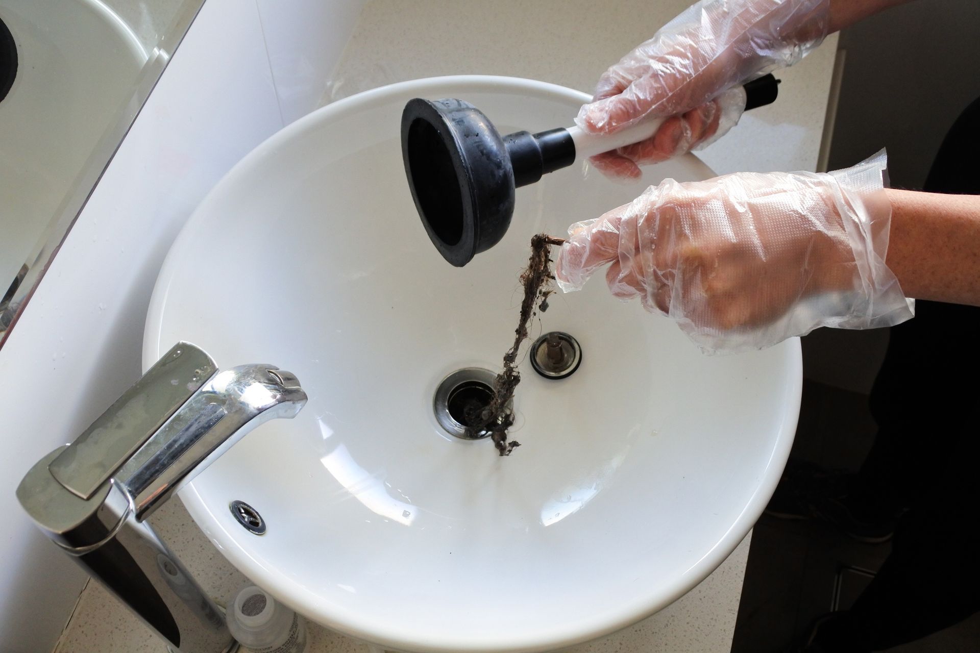 Person wearing gloves unclogging a white sink drain with a plunger, pulling out debris.