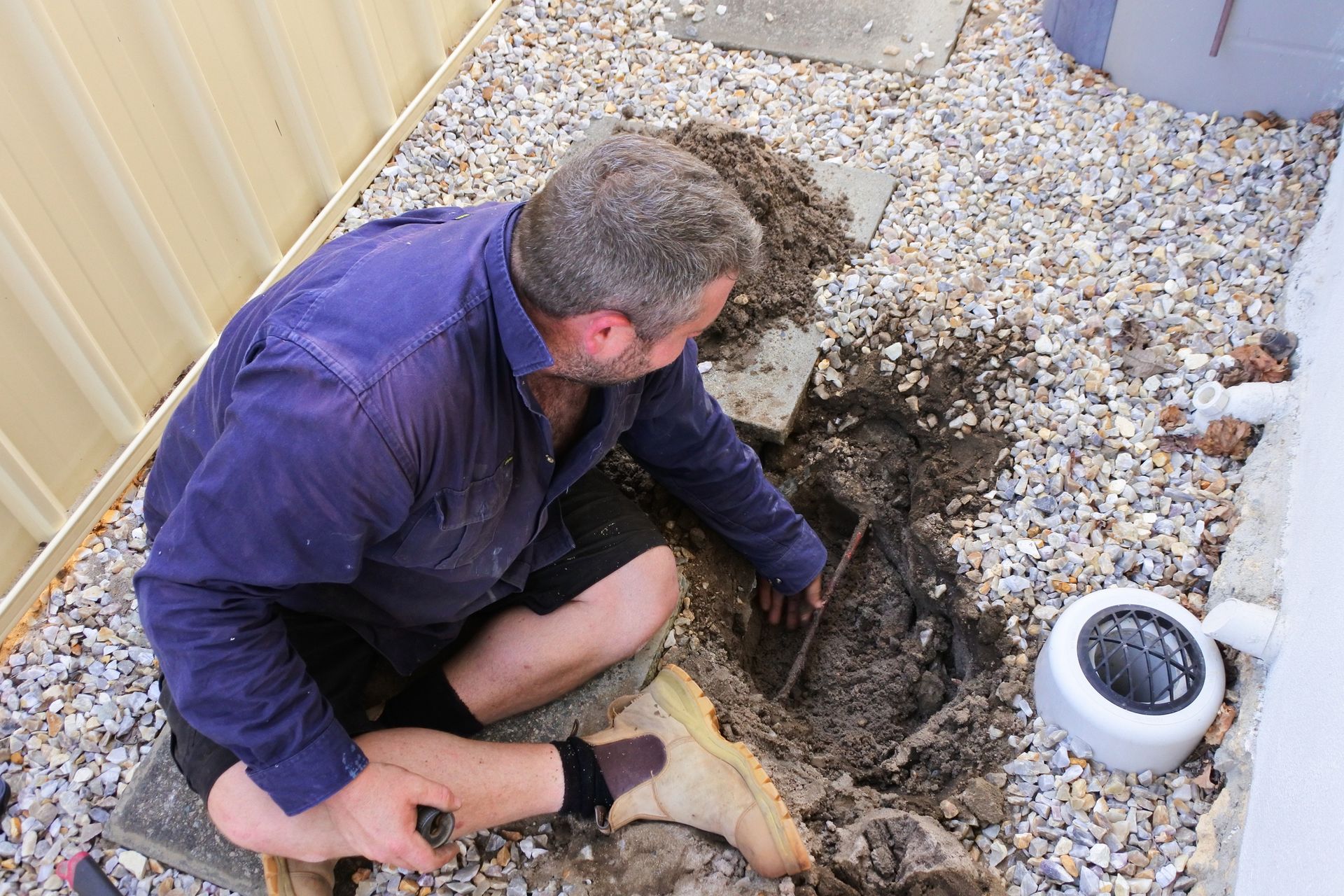 Man kneeling, digging in gravel and dirt near a white pipe in a backyard.
