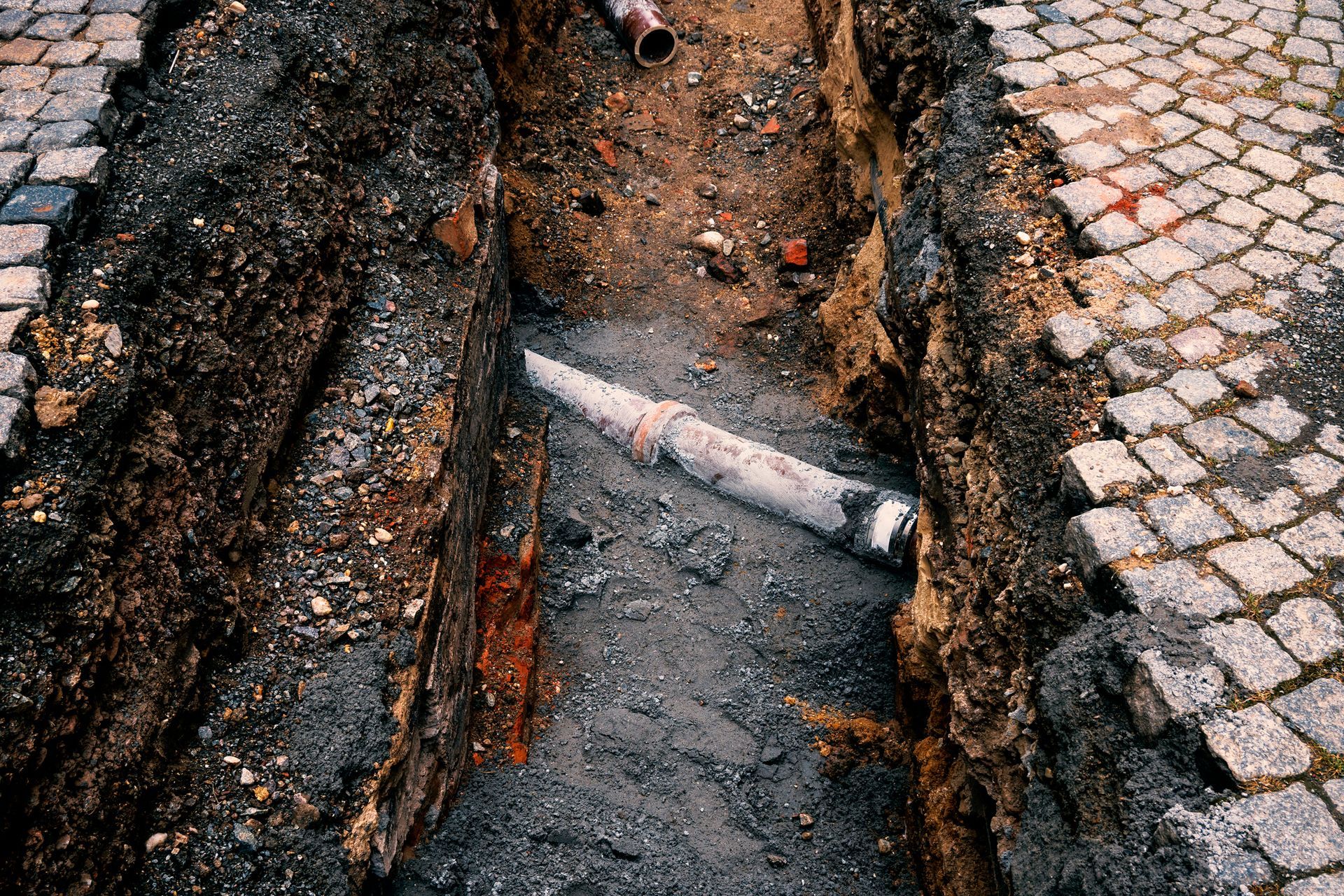 A narrow trench in a cobblestone street reveals an old pipe buried in dirt.