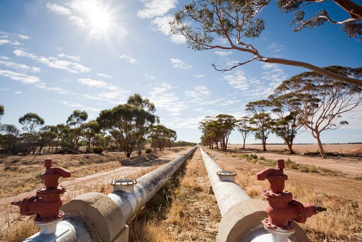 Pipes running through a dry landscape under a blue sky, trees lining either side.