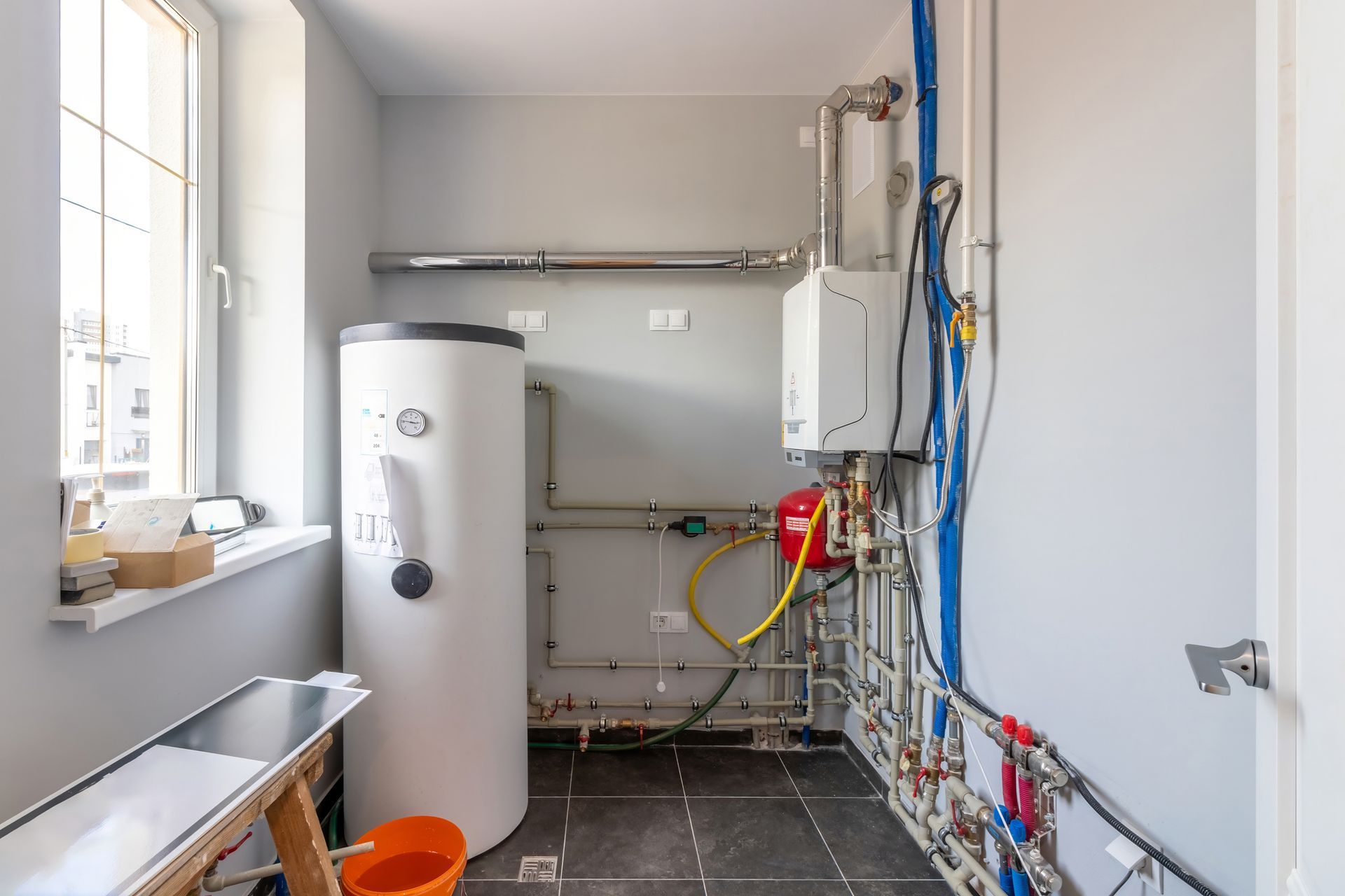 A utility room with a tall white water heater, pipes, and boiler against gray walls, with a window and a metal workbench.