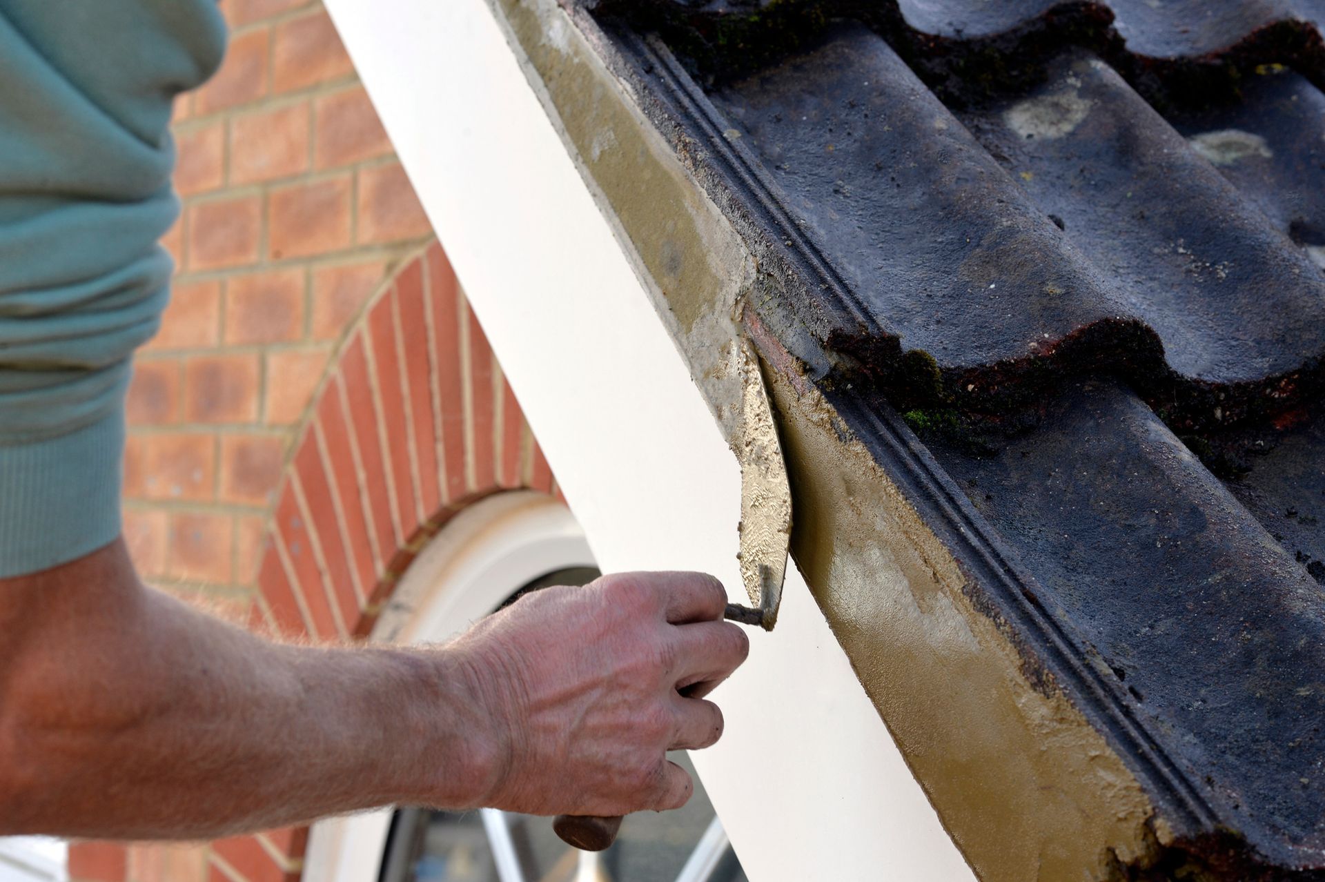 Person using a trowel to apply mortar along the edge of a roof, near dark tiles and a white-painted wall.
