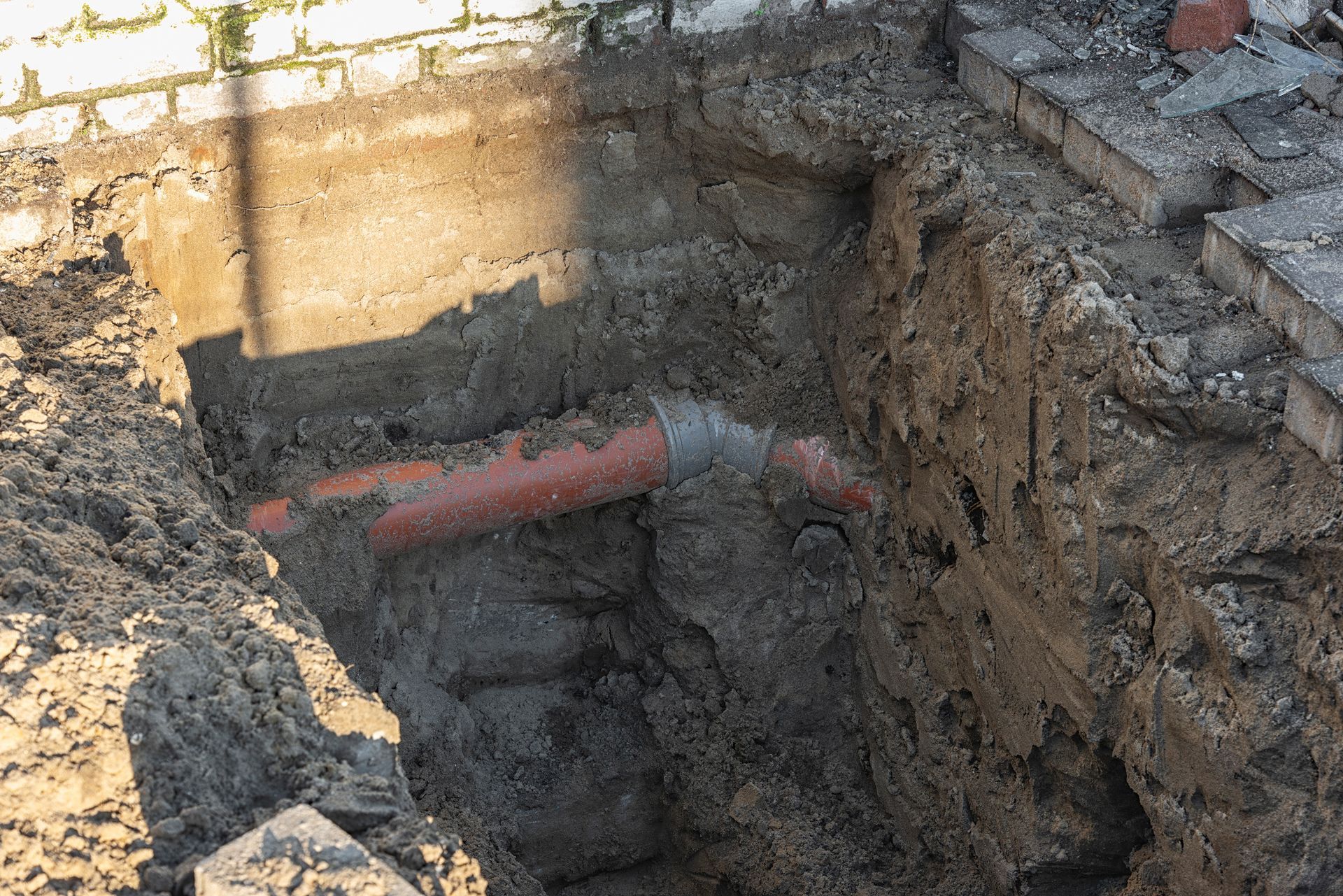A trench dug next to a brick wall, revealing orange and gray pipes in the dirt.