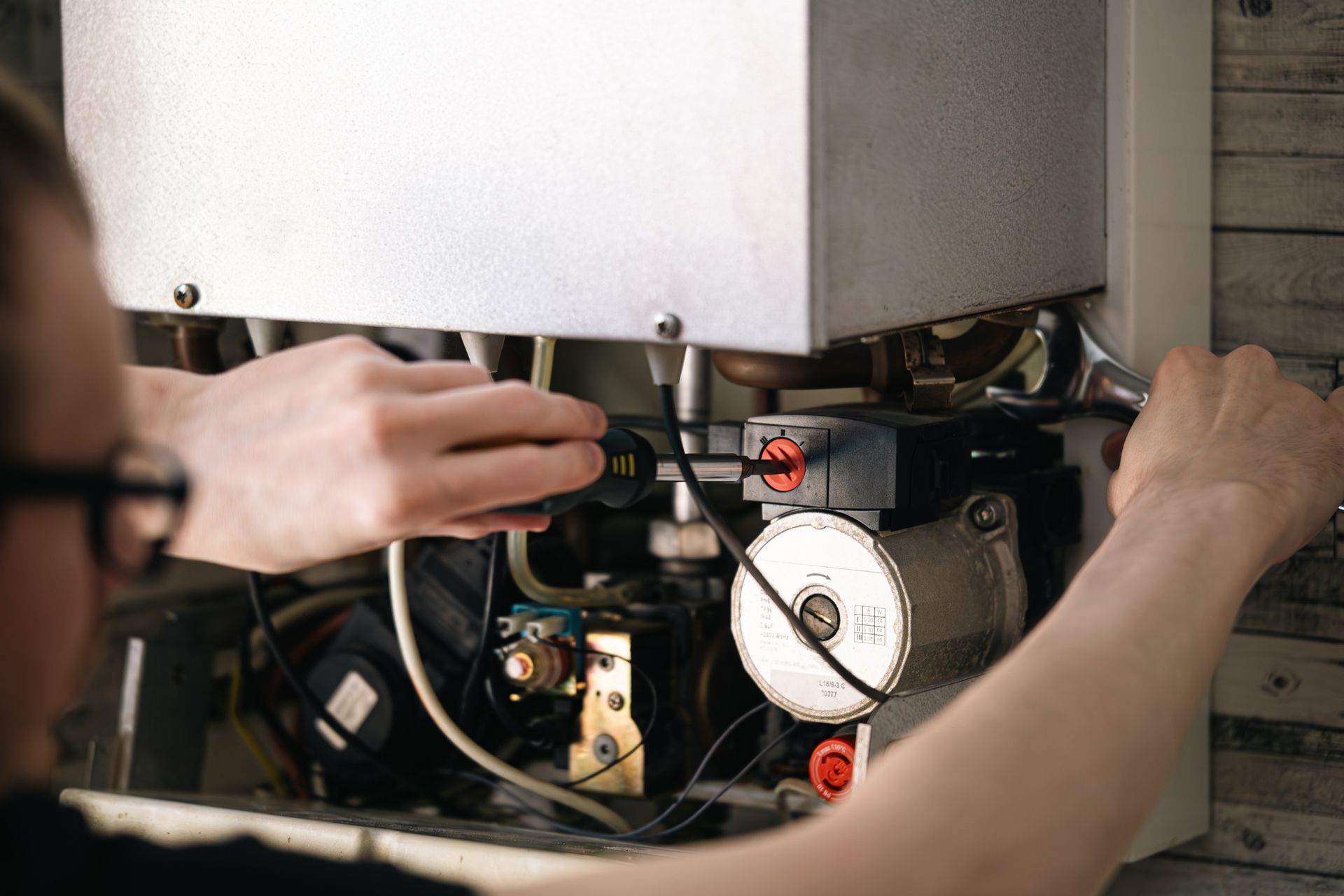 A person with glasses repairs a boiler, using a wrench and other tools.