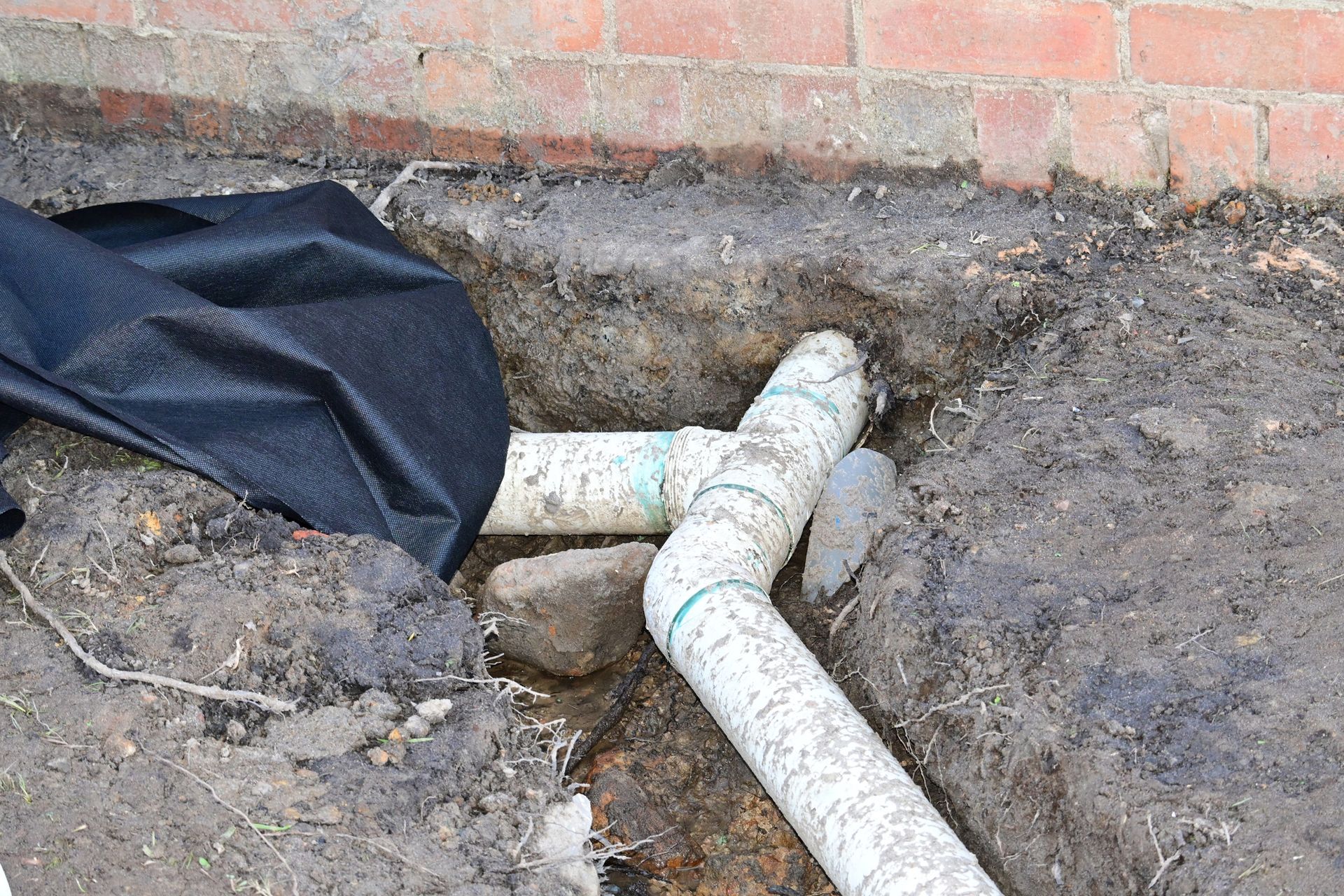 Pipes in trench near a brick wall, with landscaping fabric and dirt.