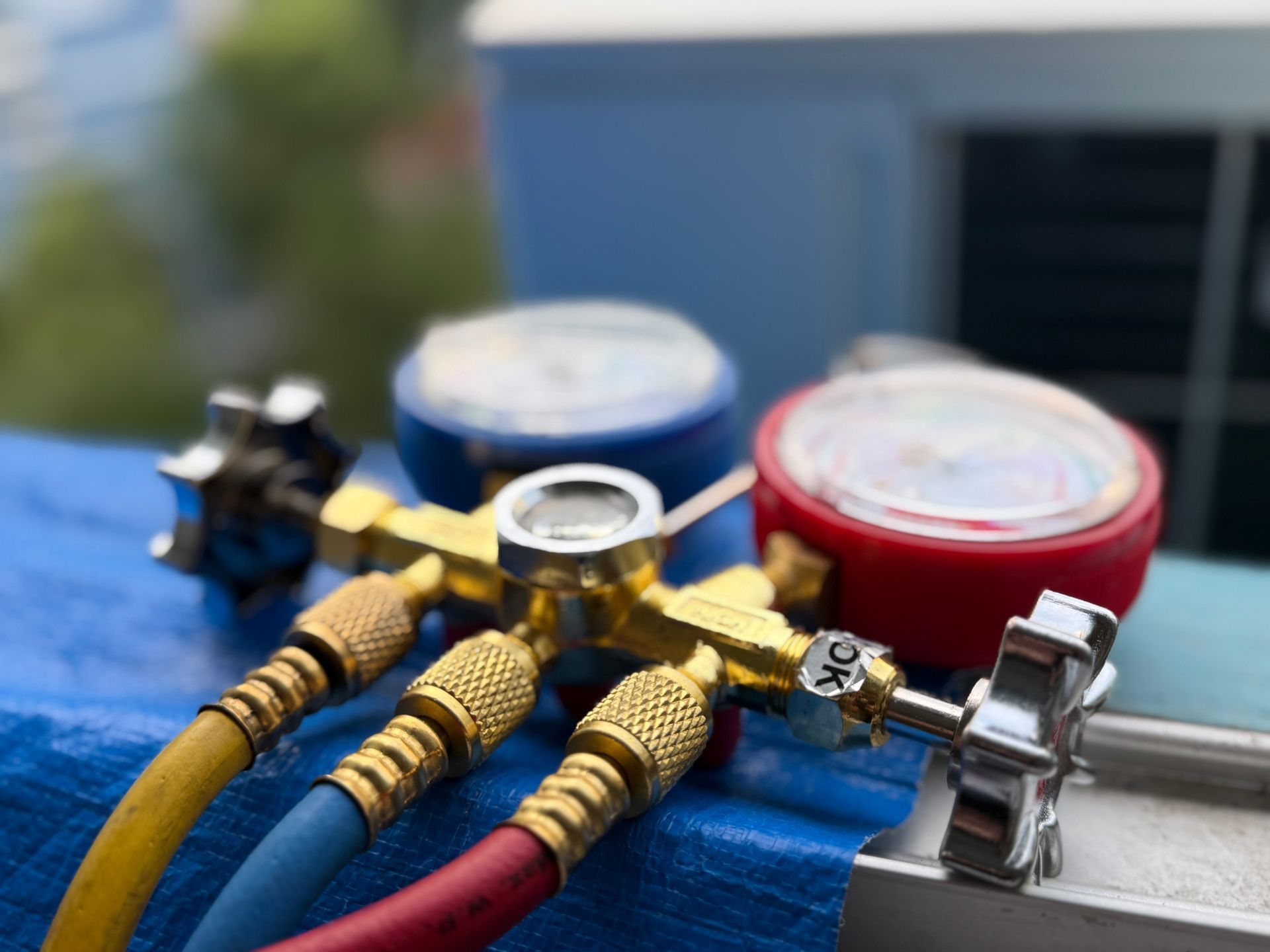 Close-up of HVAC gauges with red, blue, and yellow hoses attached, on a blue surface, with blurred background.