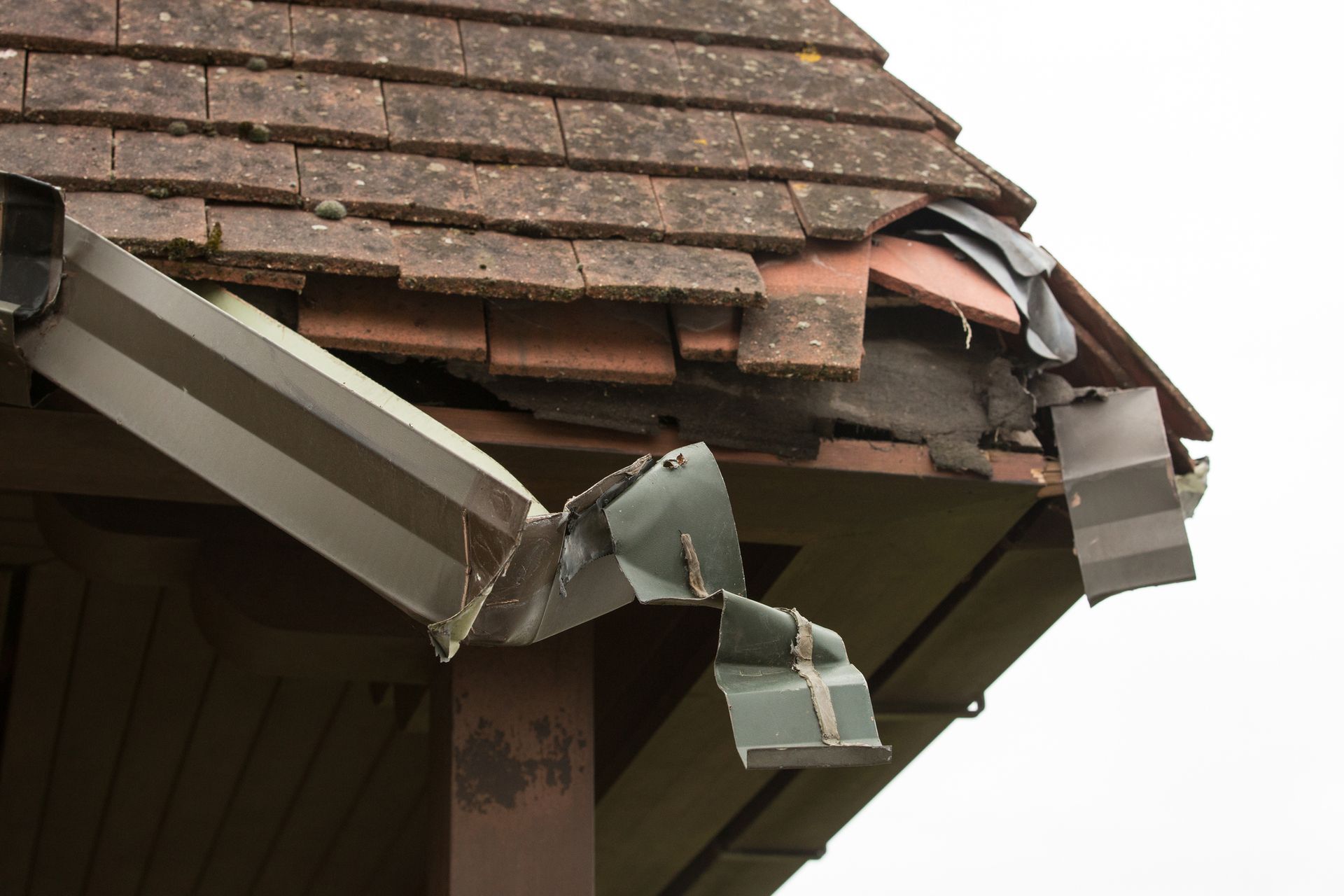 Damaged roof edge with torn metal gutter hanging down. Brown roof tiles are broken and missing.