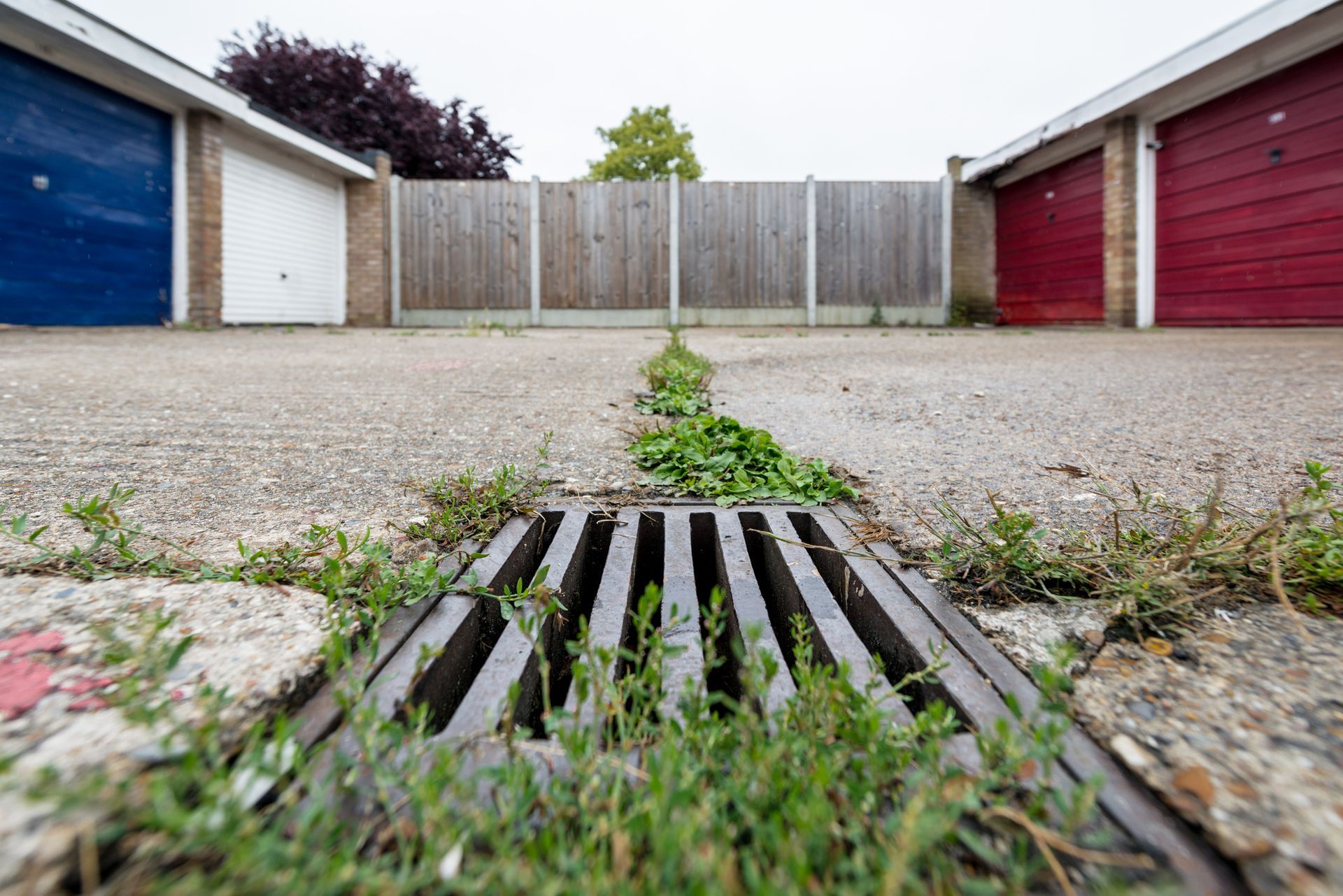 Metal drain in a concrete drive with weeds, flanked by blue, white and red garage doors and a wooden fence.