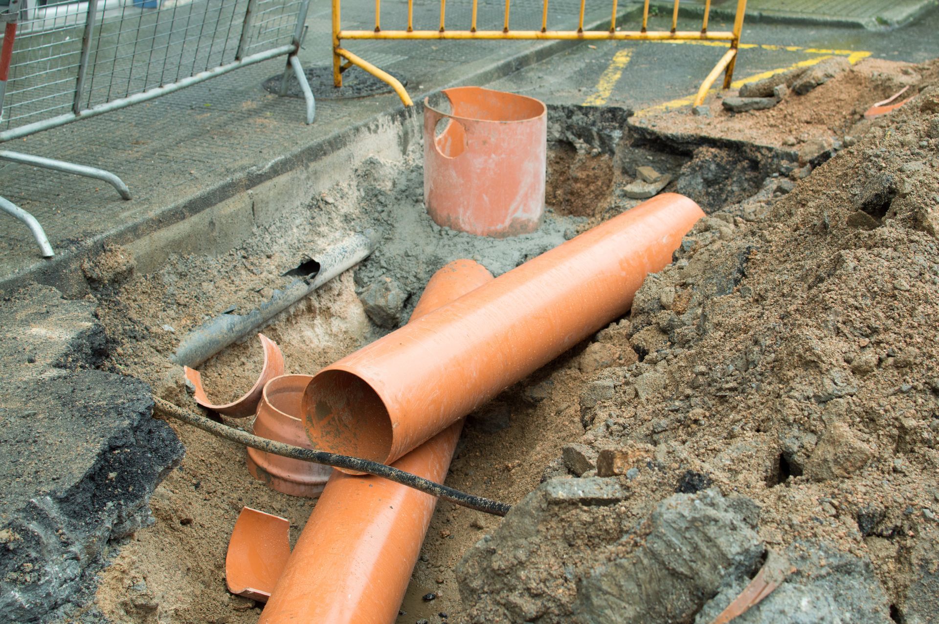 Construction site with broken orange pipes in an excavated trench near a road.