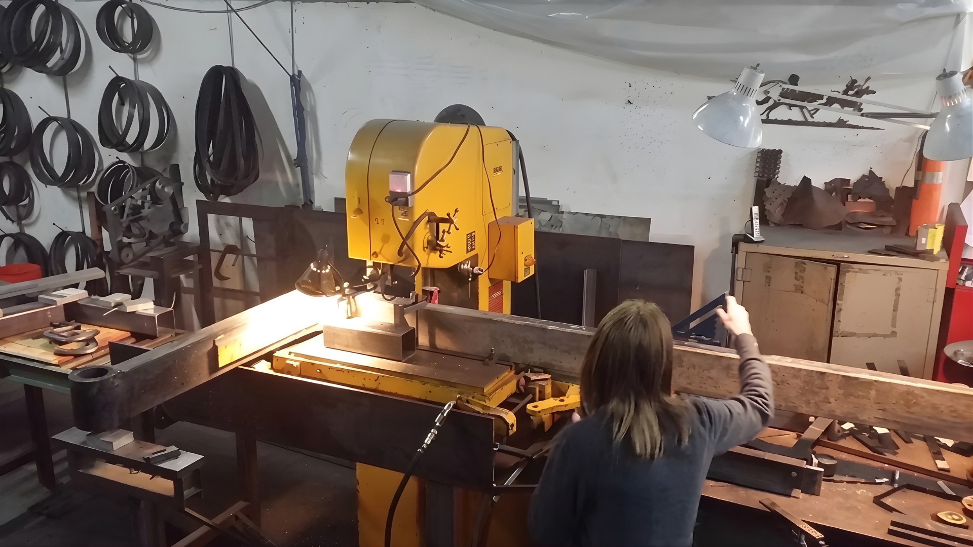 Person working at a yellow bandsaw in a cluttered workshop with tools and wood pieces around.