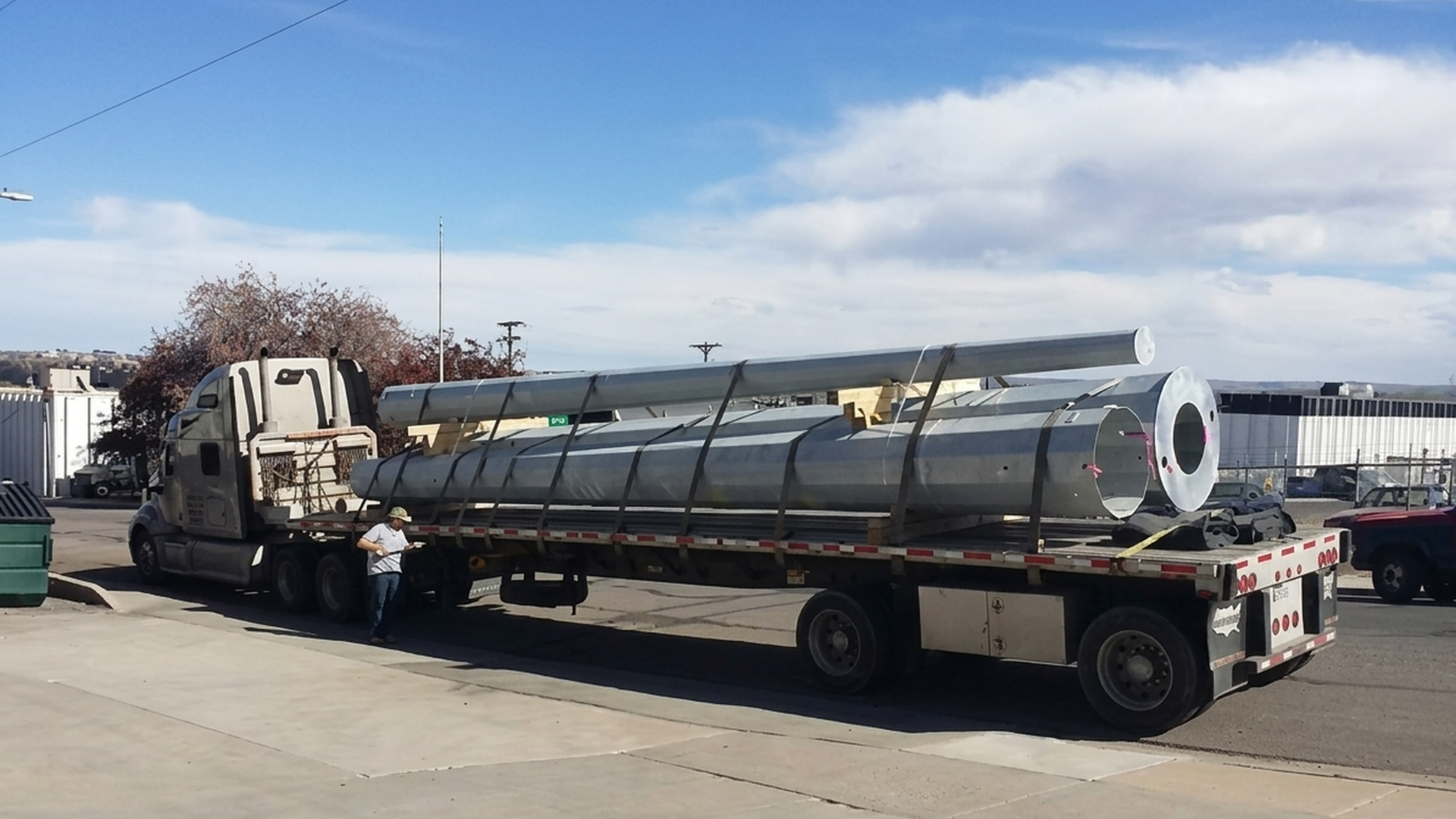 Flatbed truck hauling a large silver cylindrical tank on a roadside under a blue sky