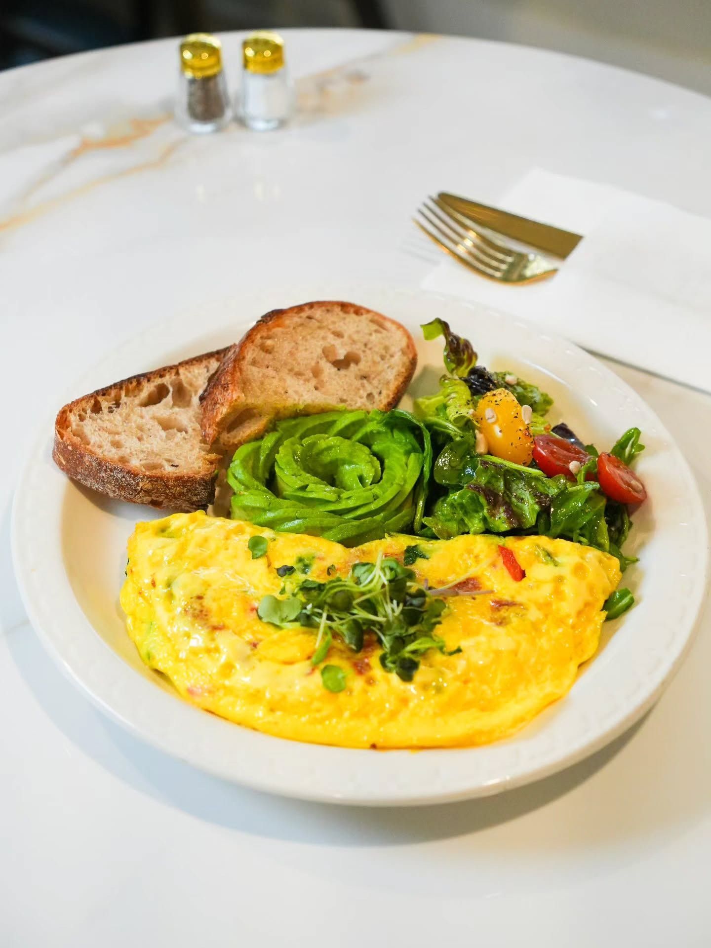 A plate featuring an omelet topped with herbs, a spiral of sliced avocado, a side salad, and two slices of toasted bread.
