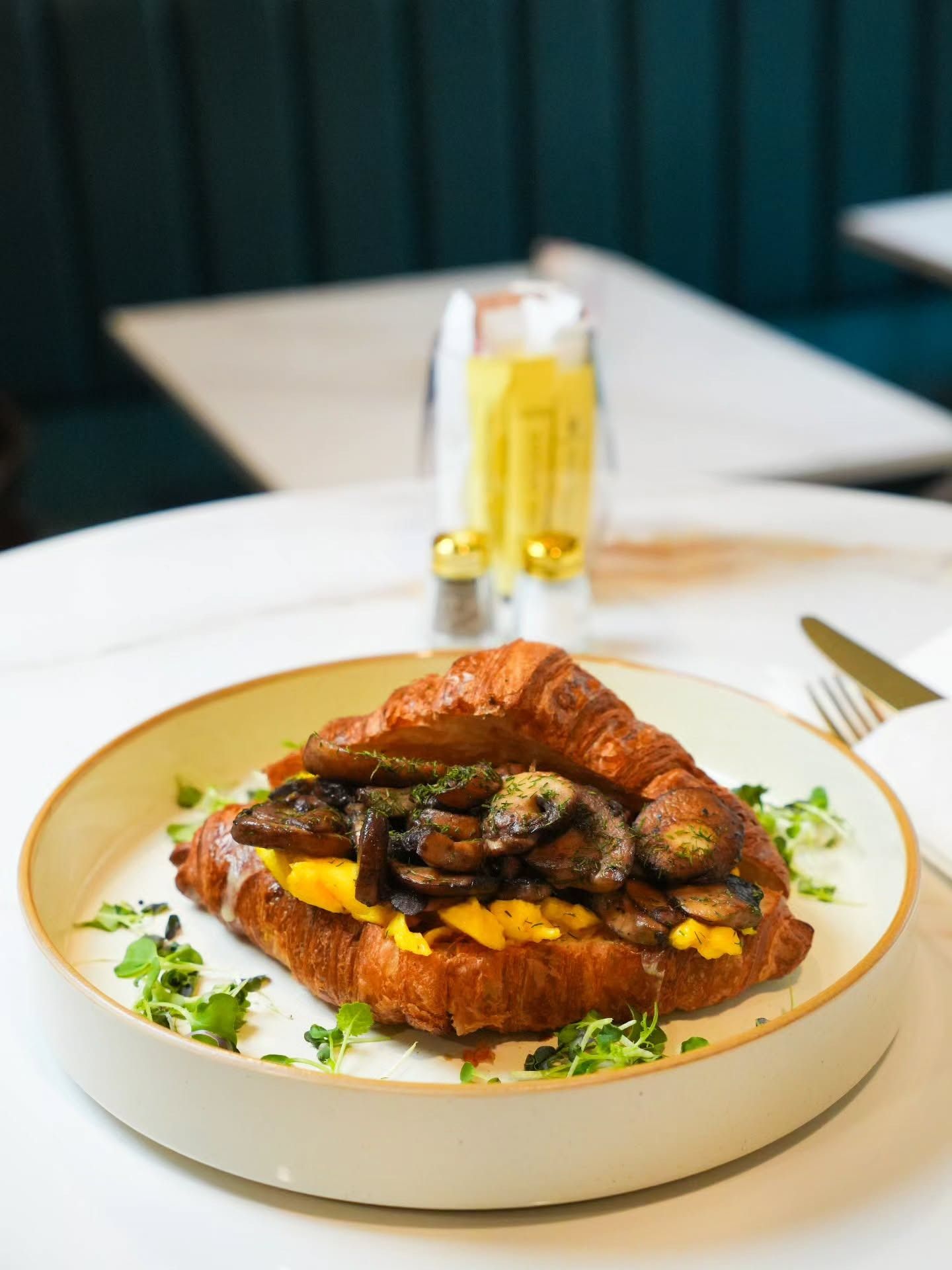 A croissant sandwich filled with scrambled eggs and sautéed mushrooms, garnished with greens on a white plate.