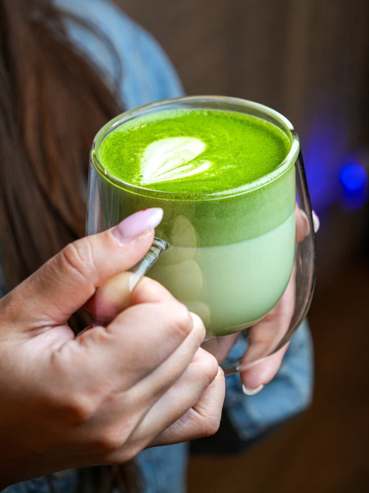 A person holding a glass mug of vibrant green matcha latte topped with a heart-shaped foam design.
