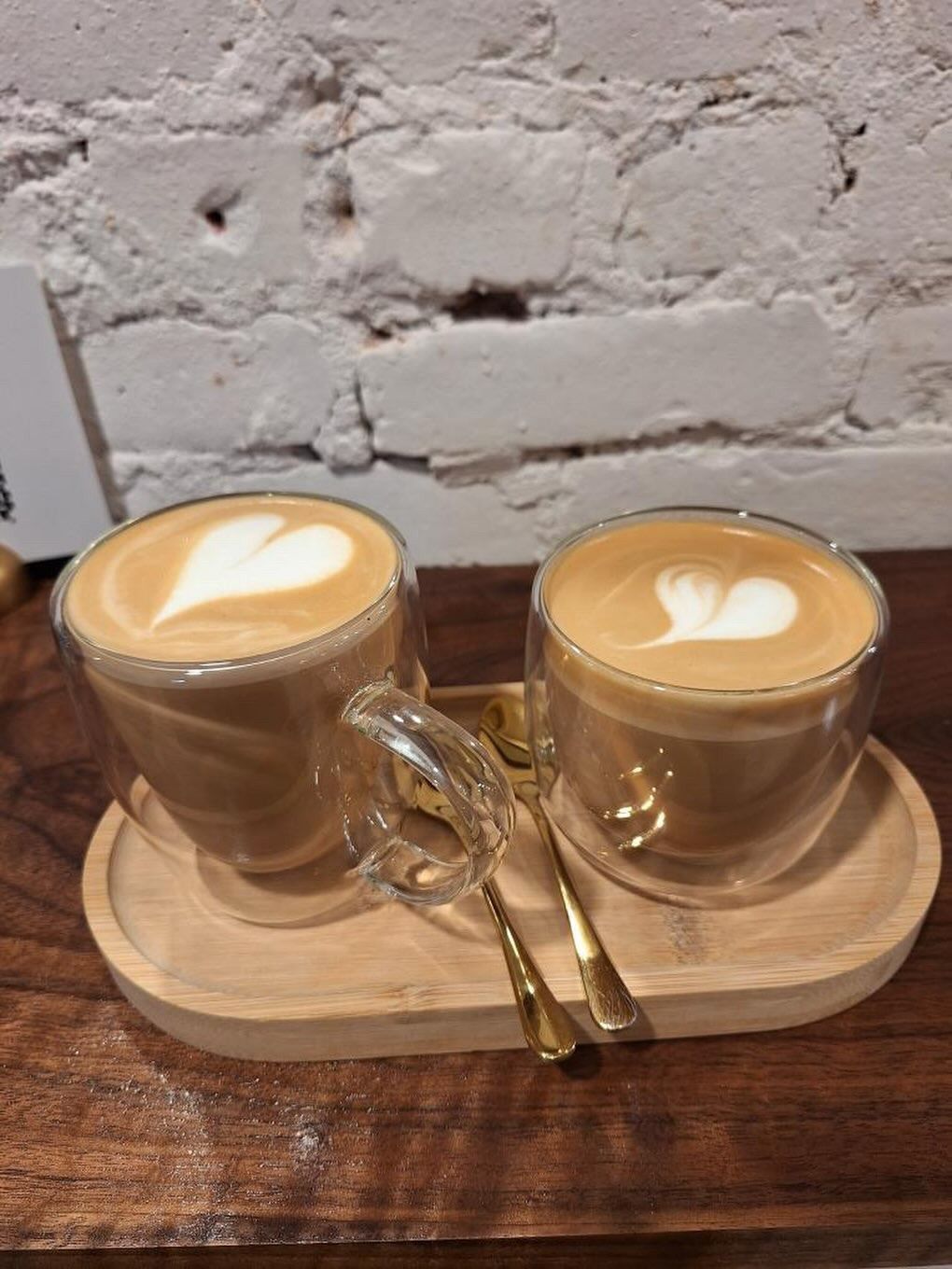 Two glasses of latte with heart-shaped foam art on a wooden tray, set against a white brick wall.