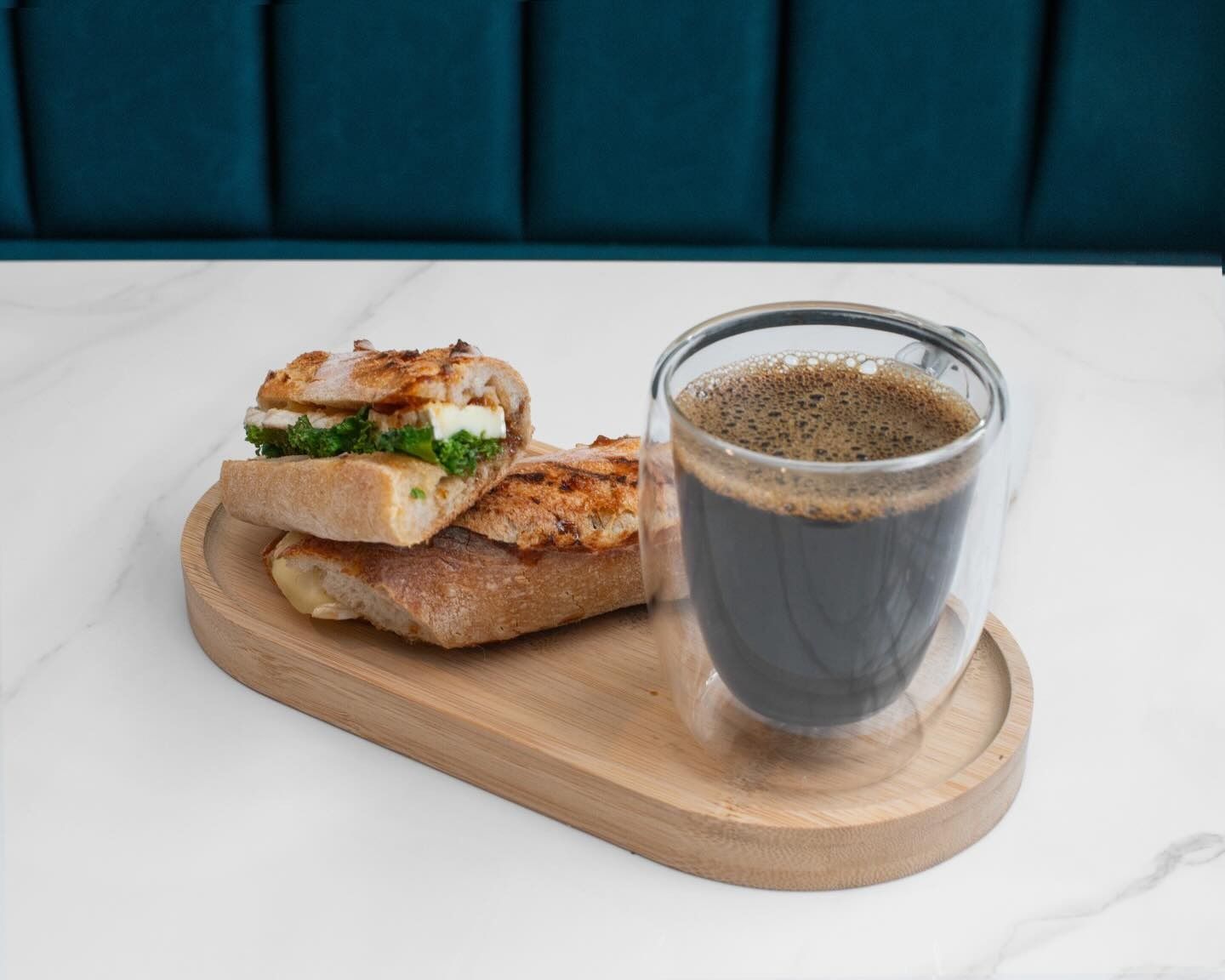 A glass of black coffee and a stacked sandwich served on an oval wooden tray atop a white table against a blue wall.