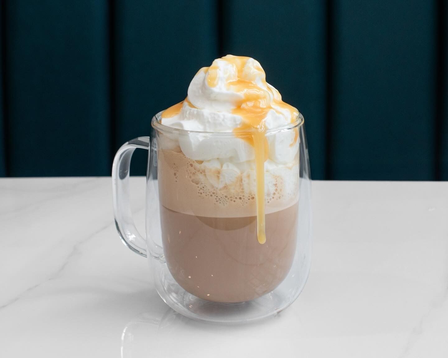 A glass mug of hot chocolate topped with whipped cream and caramel drizzle, set on a white surface against a blue wall.