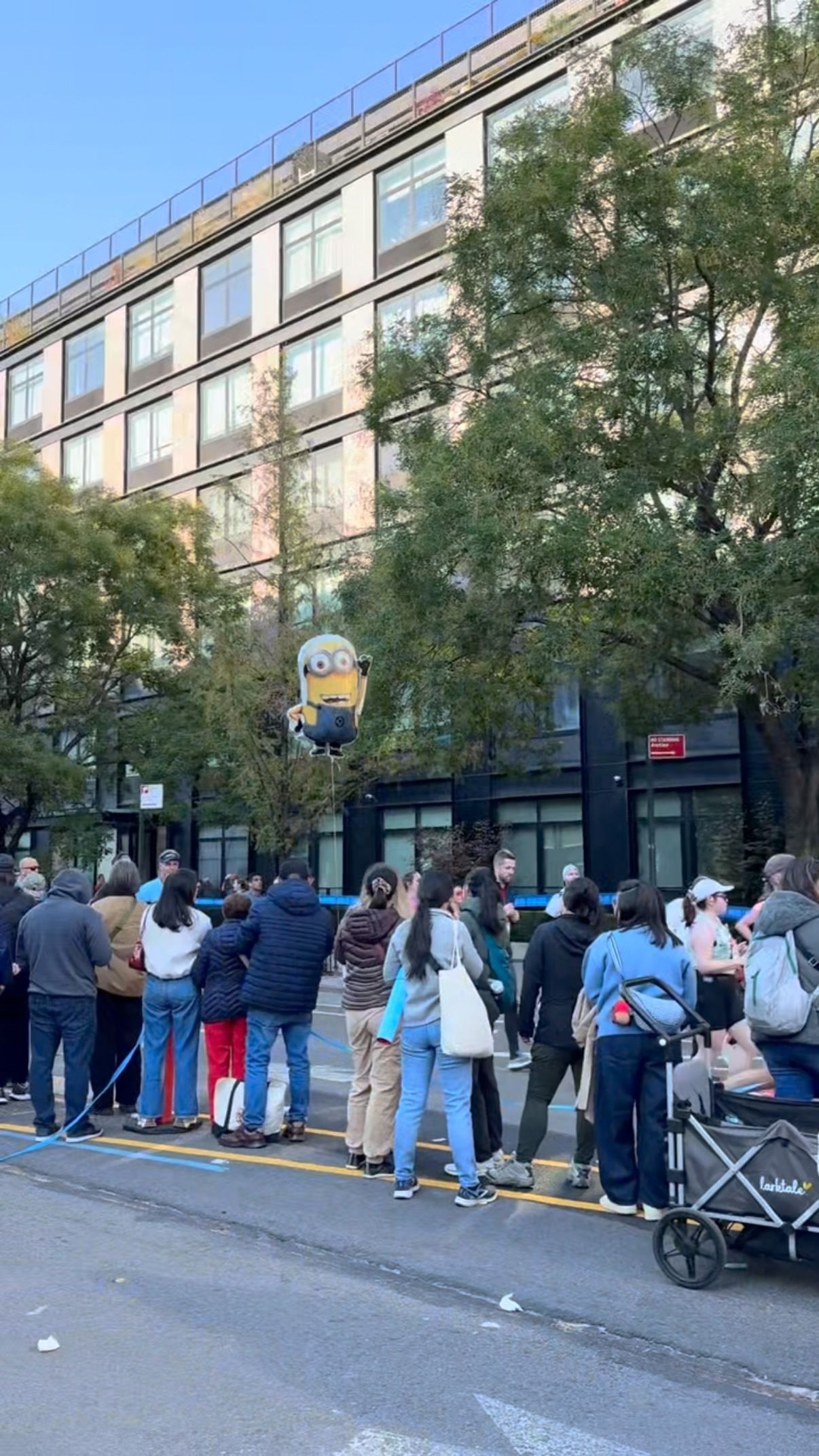 A crowd of people stands on a city street watching a large Minion character balloon floating in the air.