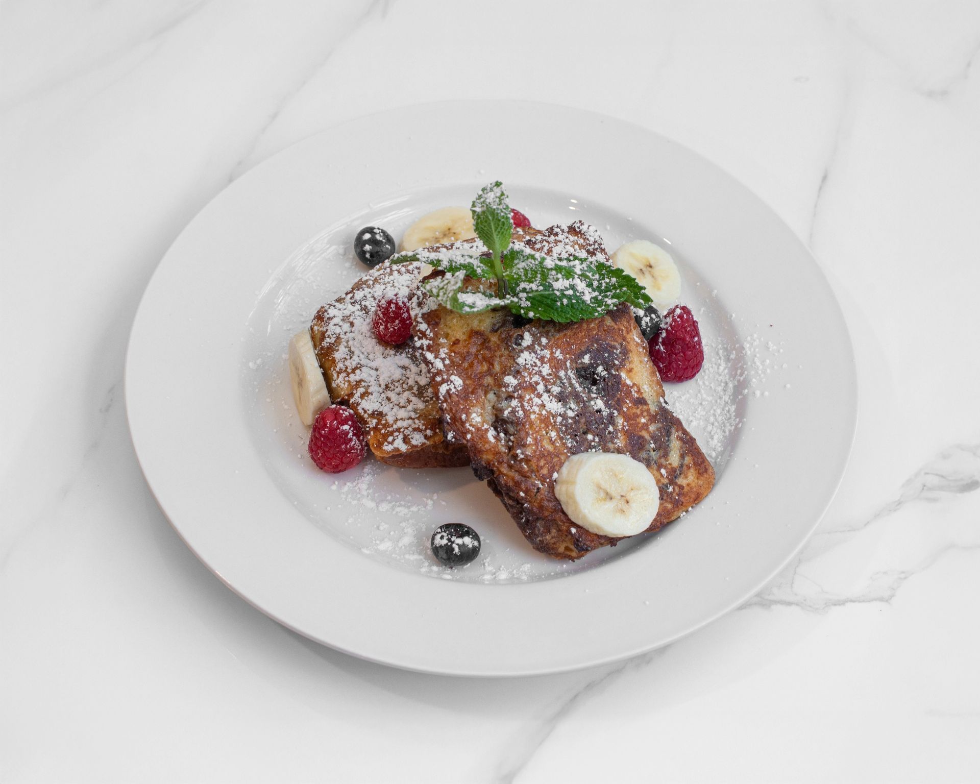 Two slices of French toast topped with fresh raspberries, blueberries, banana slices, and mint on a white plate.