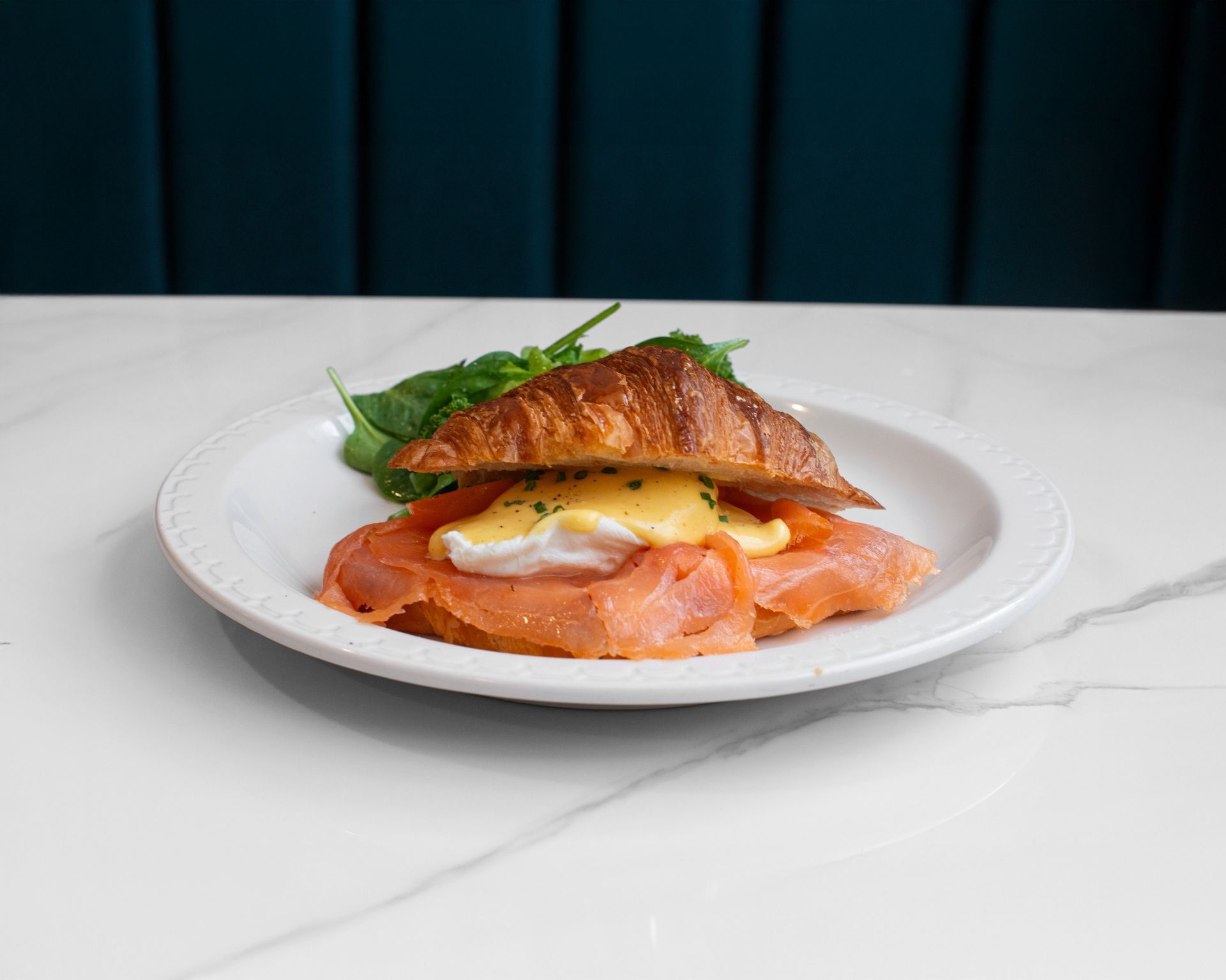 A croissant sandwich with smoked salmon and a poached egg topped with hollandaise sauce on a white plate with greens.