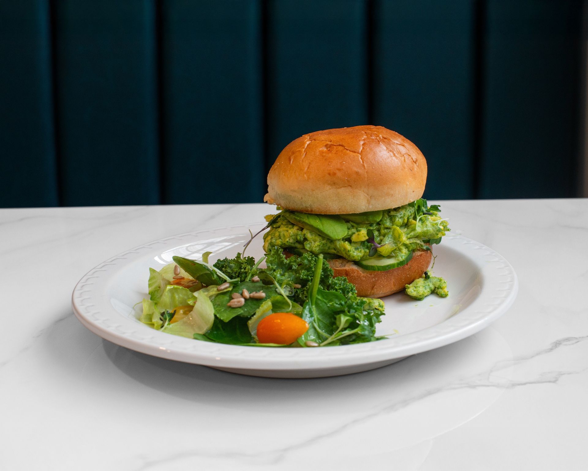 A burger with avocado and a side salad on a white plate against a dark teal background.