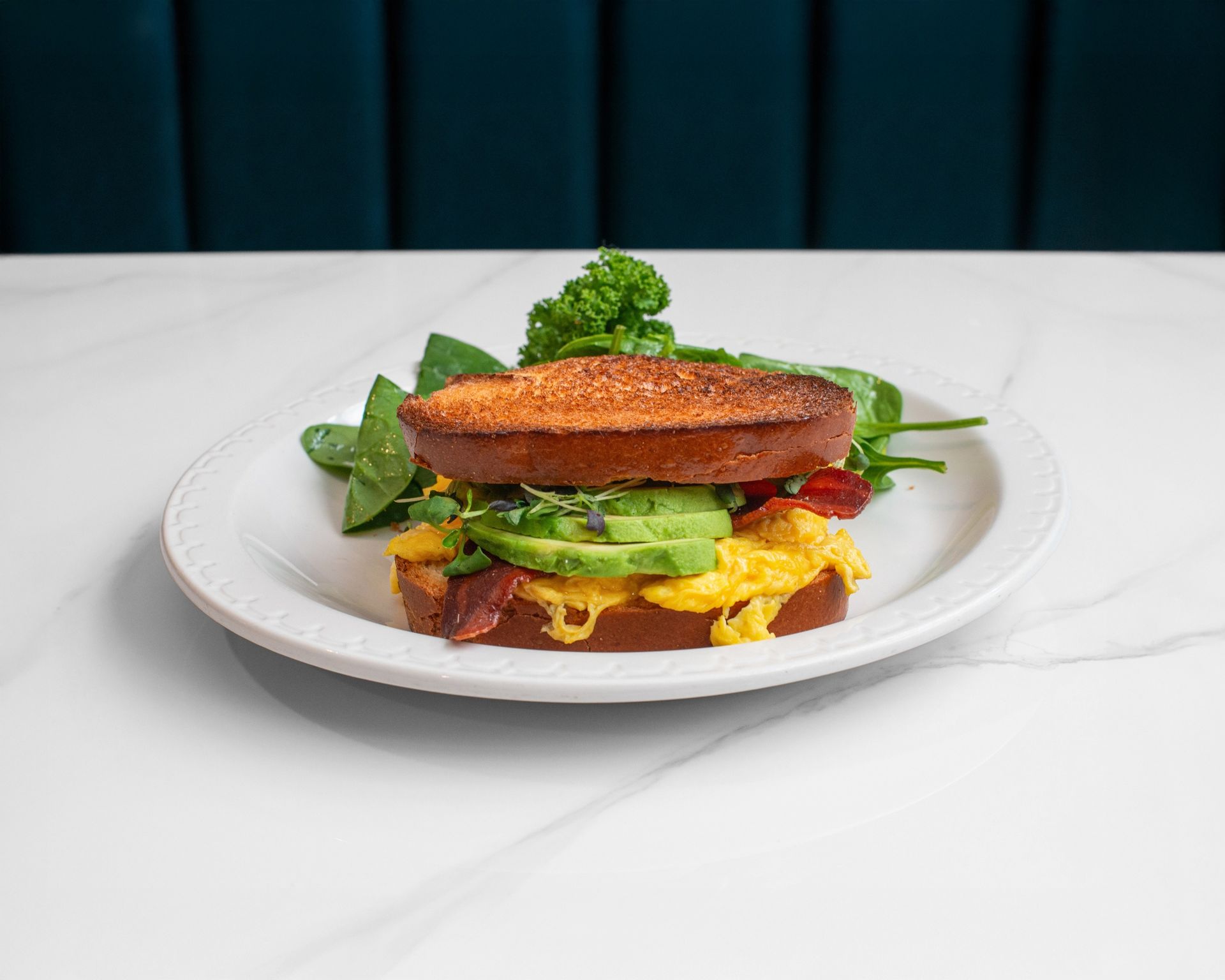 A breakfast sandwich with scrambled eggs, avocado, and bacon on toasted bread, served with a side of greens on a white plate.