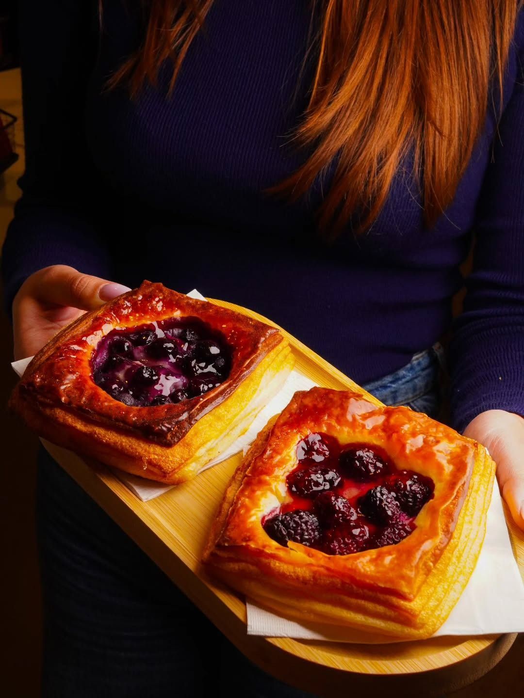 A person in a blue sweater holds a wooden tray with two square berry pastries on white napkins.