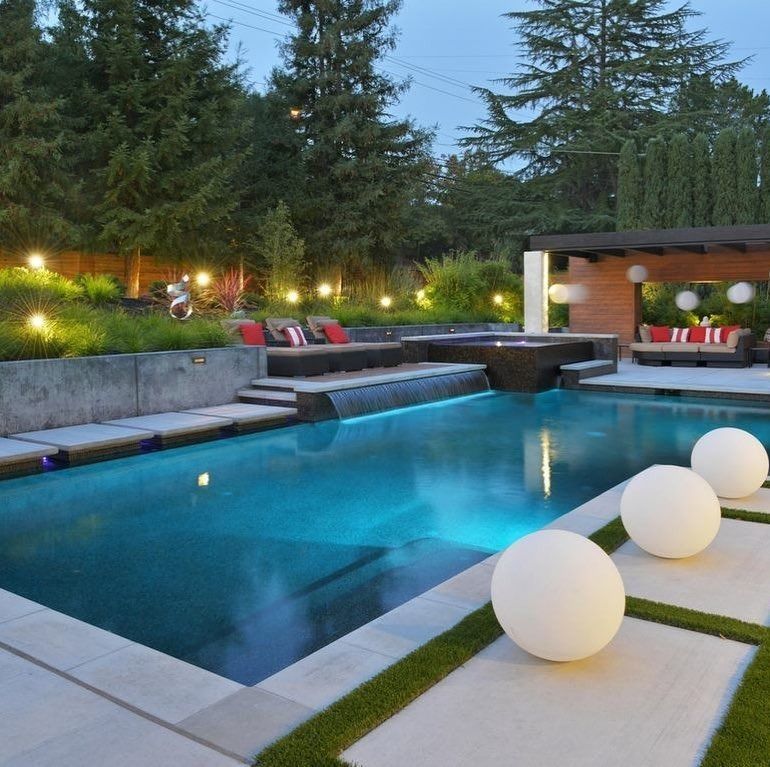 Modern backyard pool at dusk with glowing lights, lounge seating, and white spherical lights along the deck