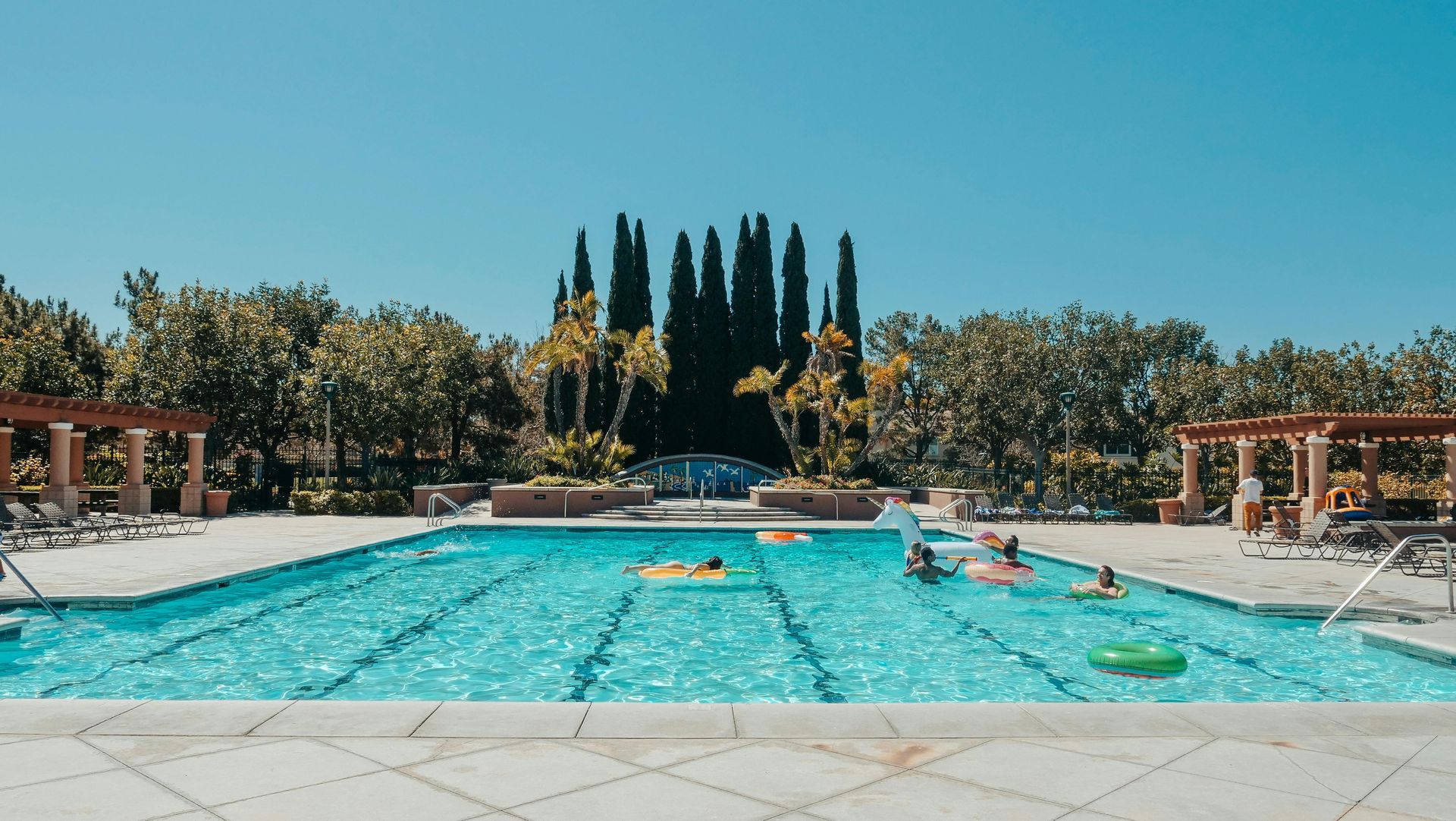 Outdoor swimming pool with turquoise water, lounge chairs, and tall trees under a clear blue sky