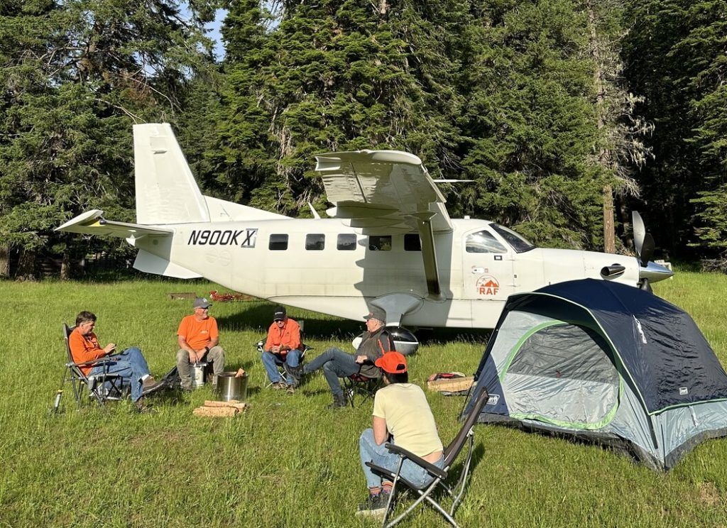 A group of people are sitting around a campfire in front of a small plane.