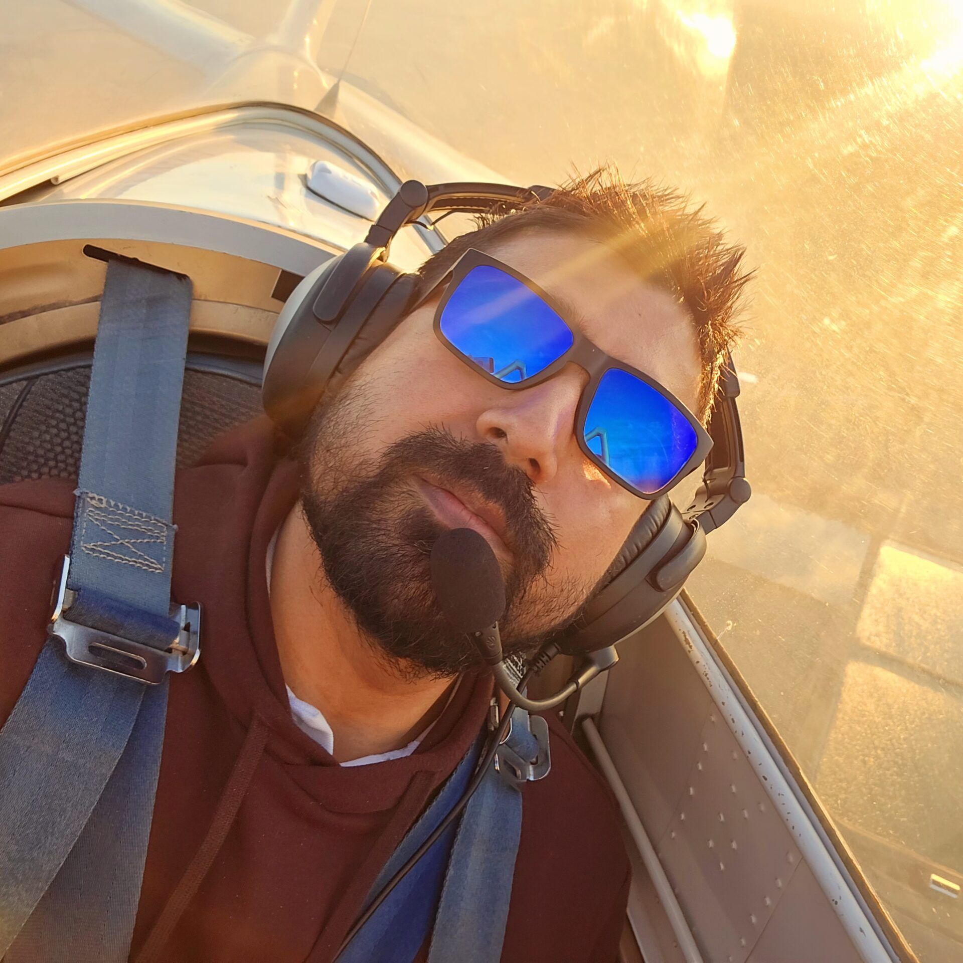 Man wearing sunglasses and headset in a cockpit, illuminated by warm sunlight.