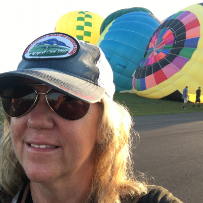 A woman wearing a hat and sunglasses stands in front of hot air balloons