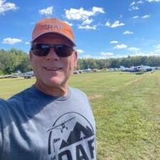 A man wearing sunglasses and a hat is taking a selfie in a field.