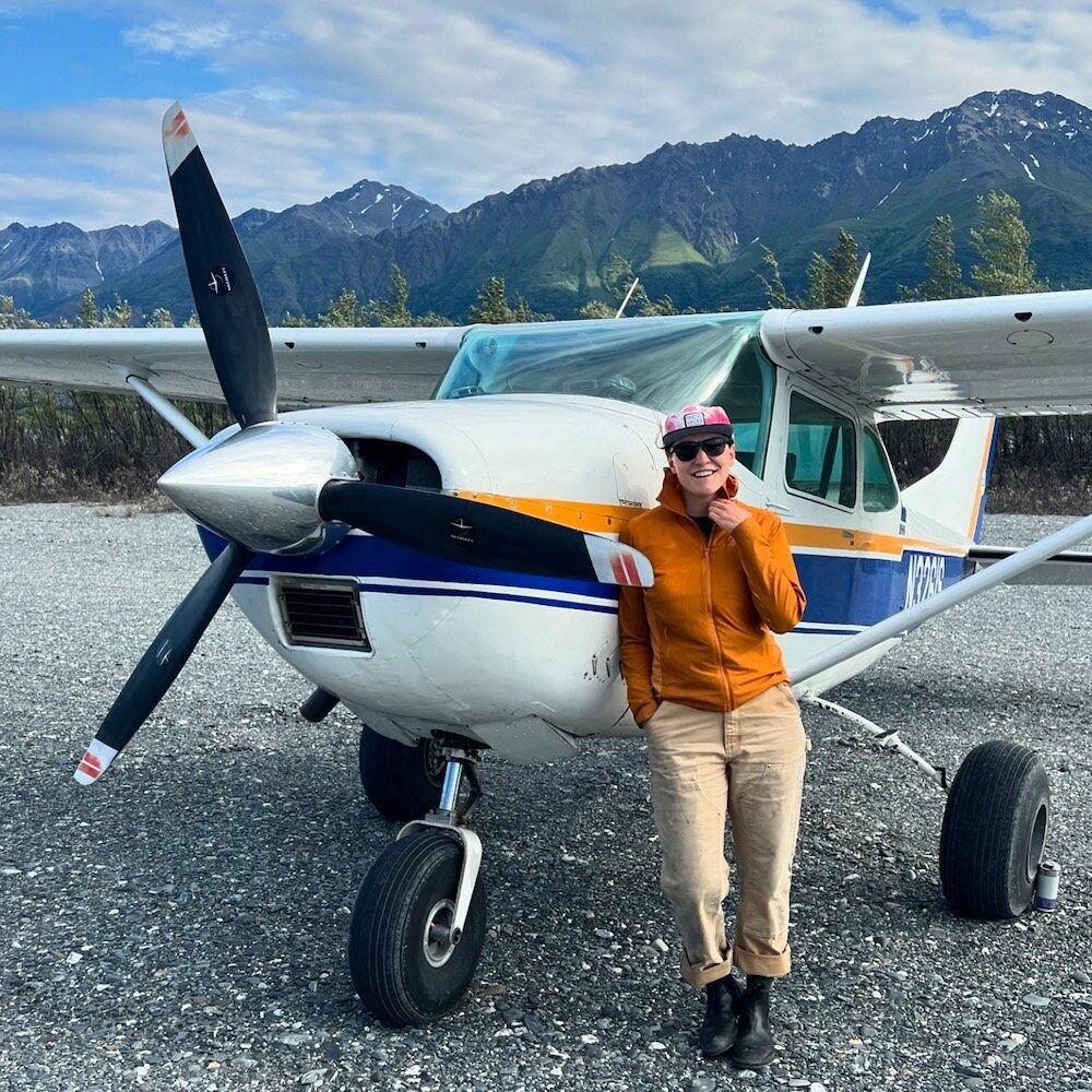 A man is standing in front of a small plane with mountains in the background